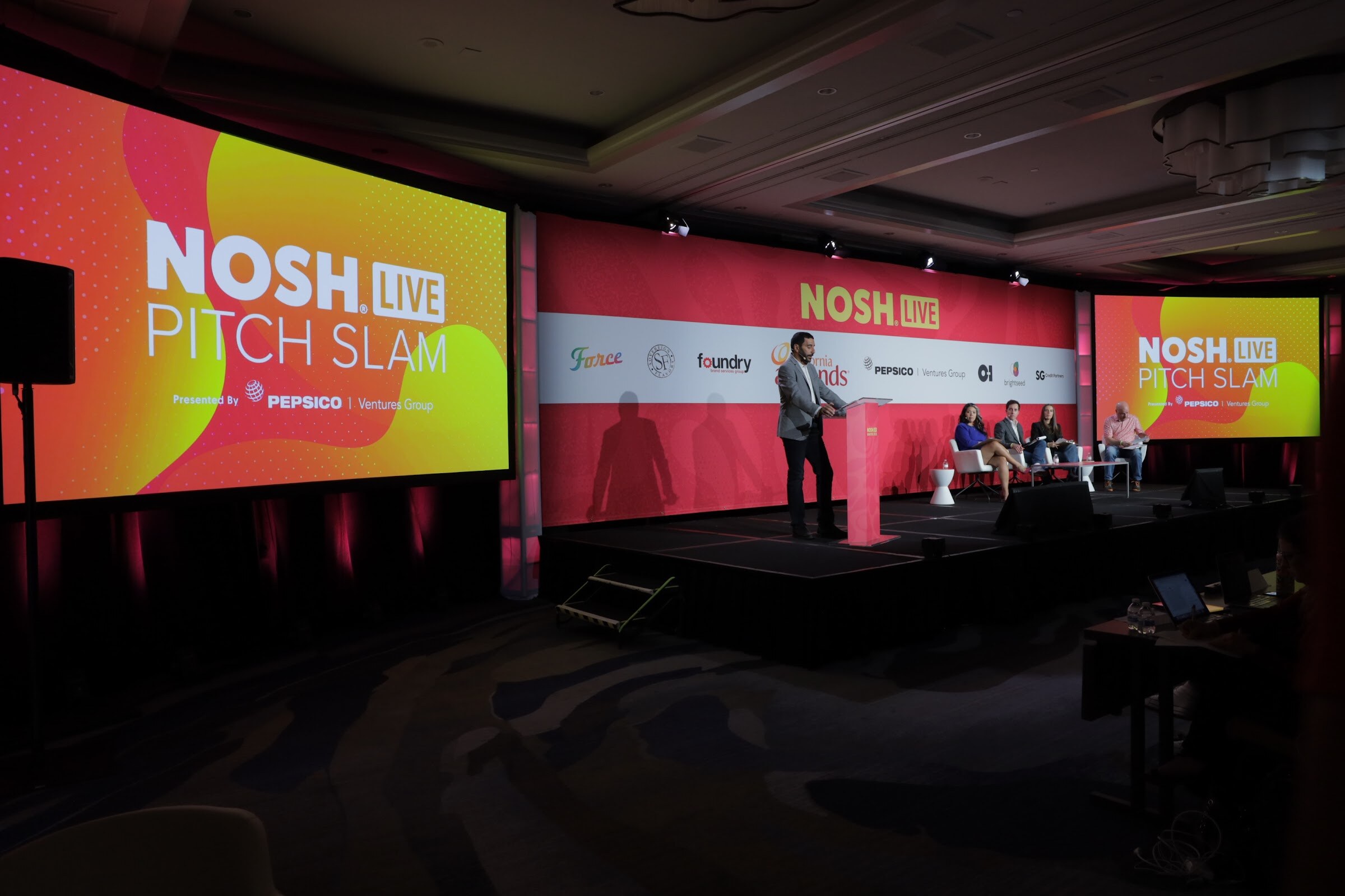 A man at a podium on stage with a large orange NOSH LIVE Pitch Slam title slide behind him.
