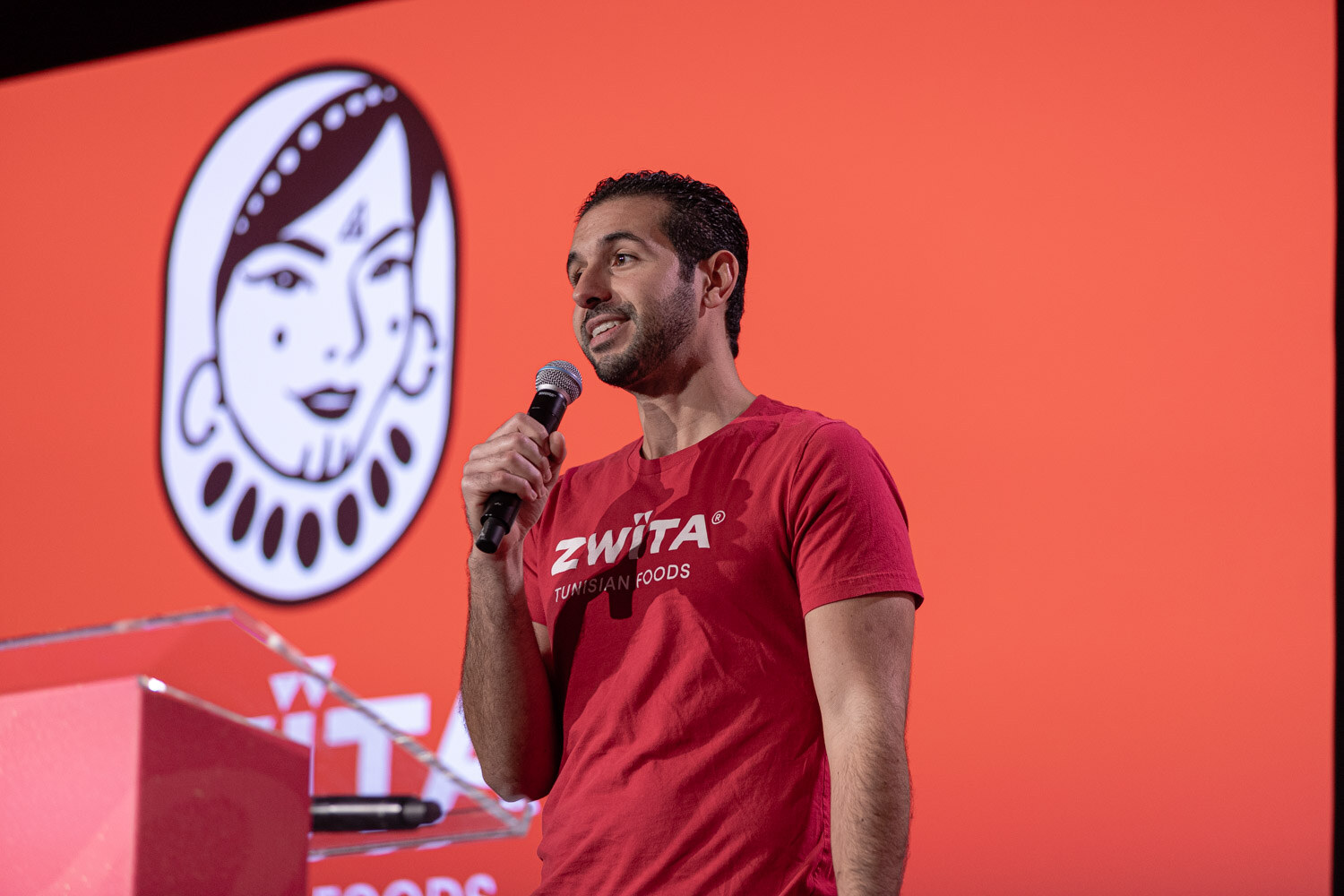 A man in a red Zwita Tunisian Foods shirt speaking into a microphone in front of an orange slide.