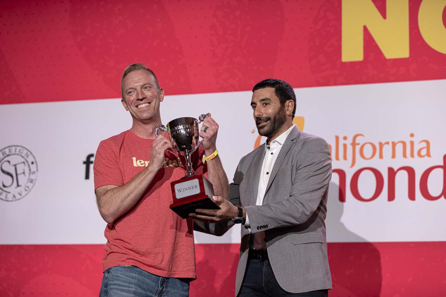 A man in a red Lentiful shirt and a man in a gray blazer holding a silver trophy marked Winner.