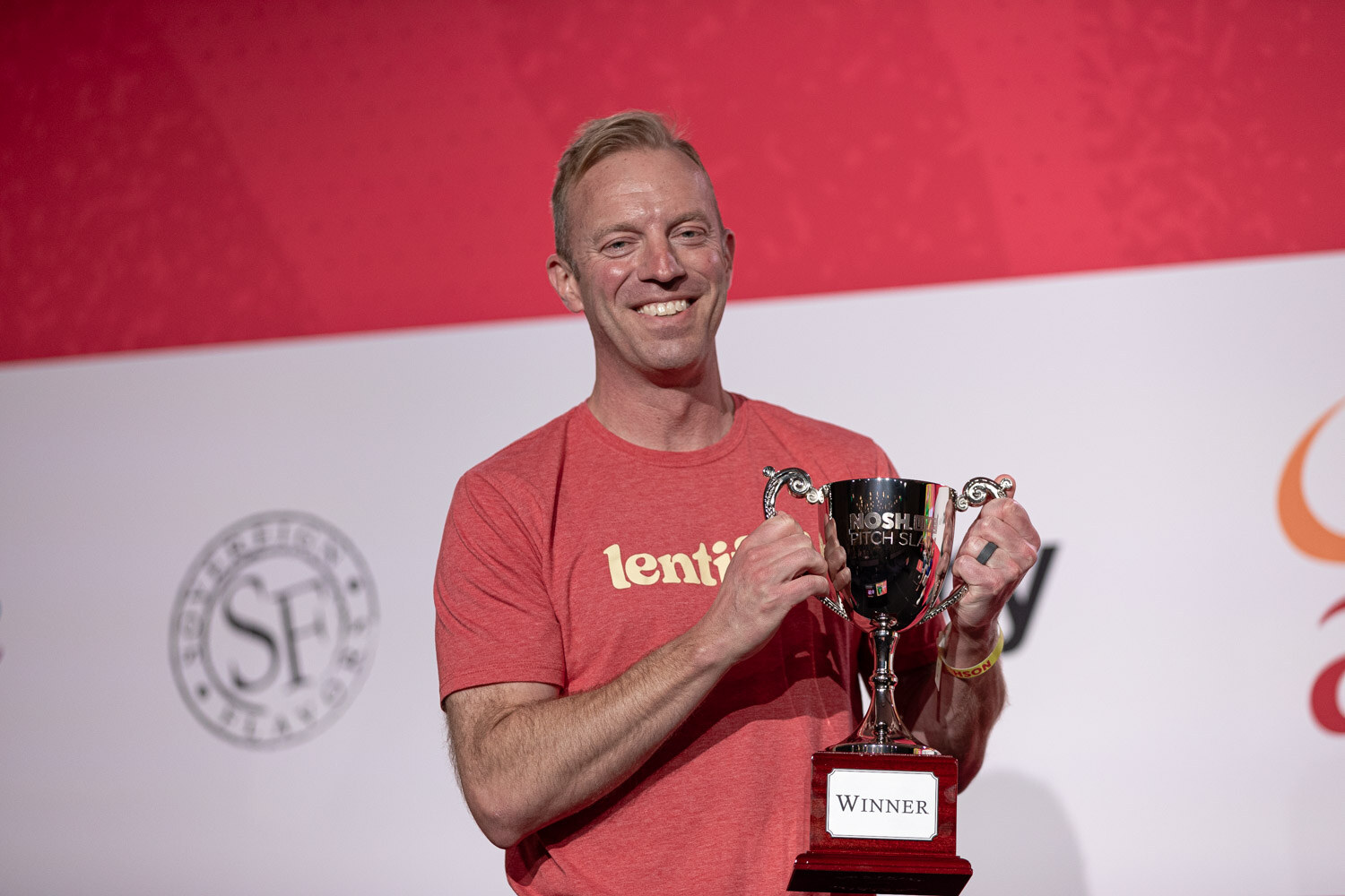 A smiling man in a red Lentiful shirt holding a trophy with a Winner plate.
