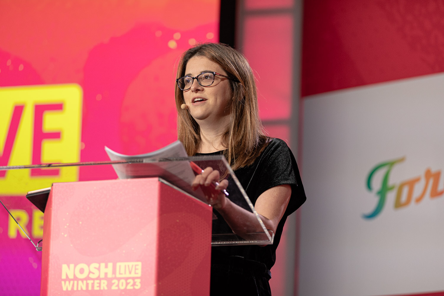 A woman speaking at a podium in front of a pink NOSH LIVE screen.