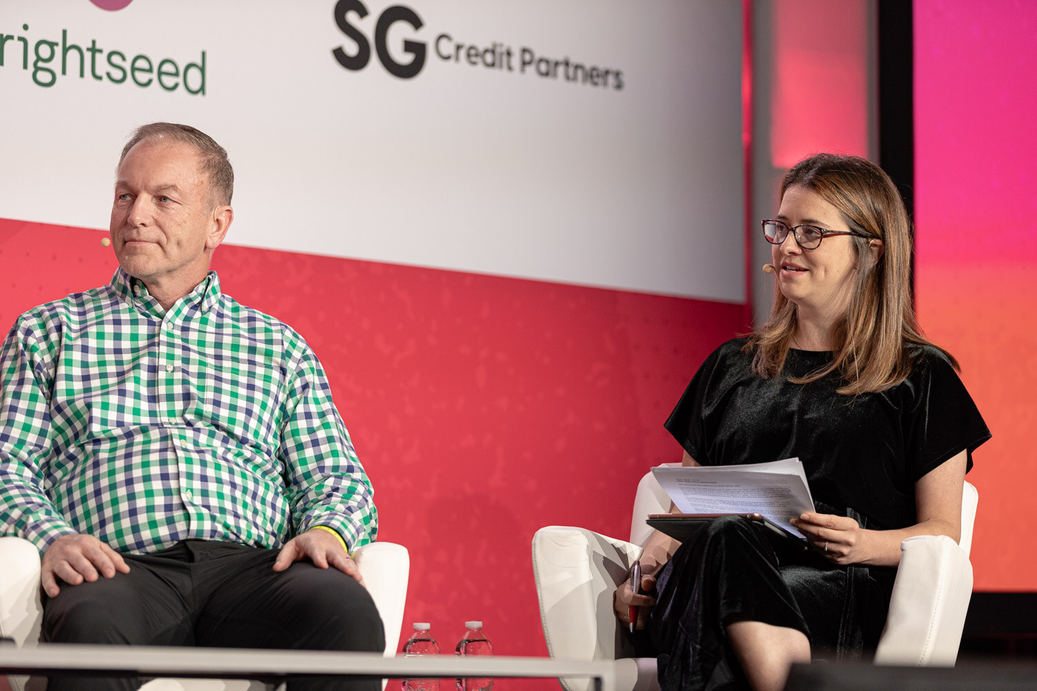 A man in a green plaid shirt and a woman with papers seated on stage.