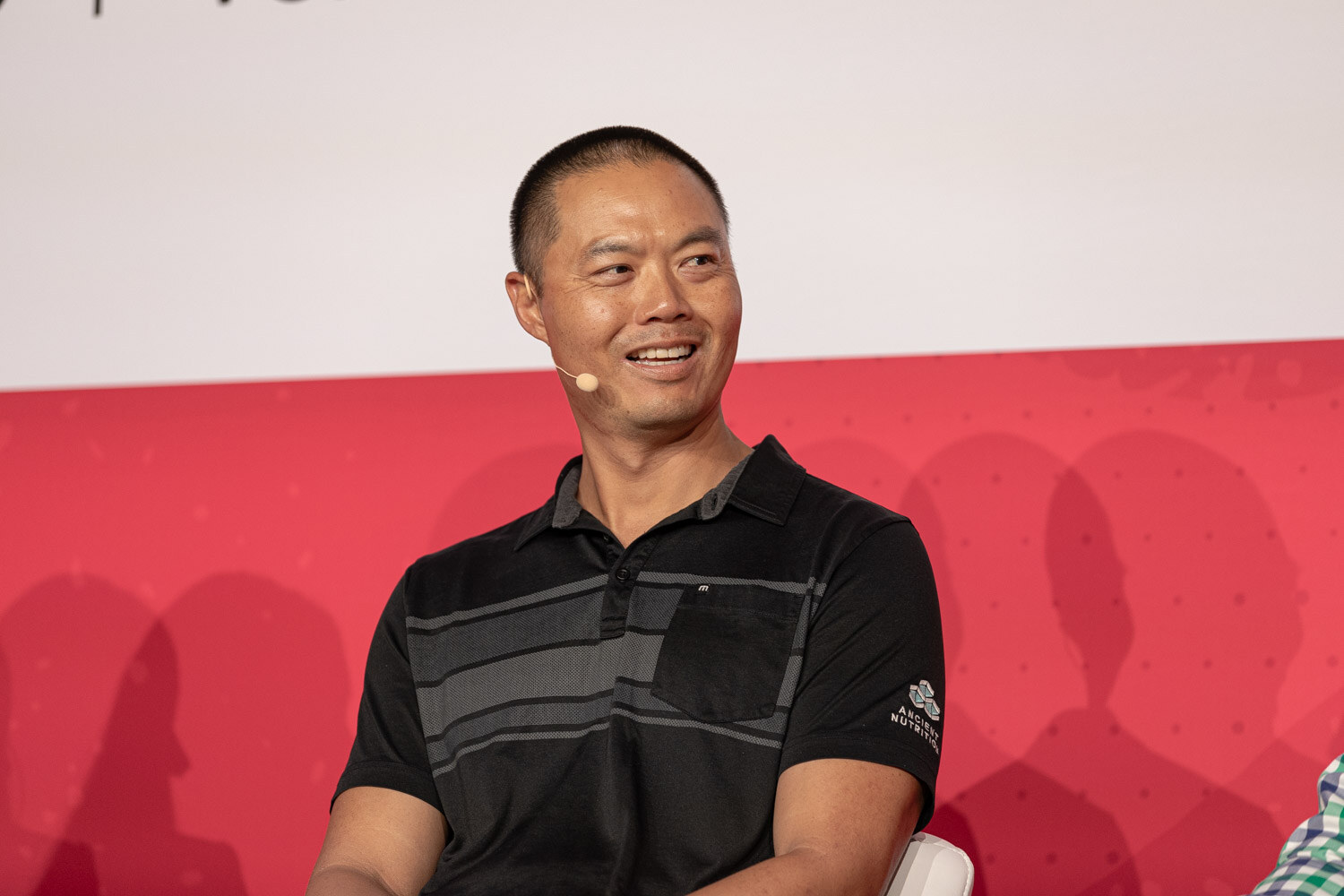A man in a black polo smiling while seated on stage.
