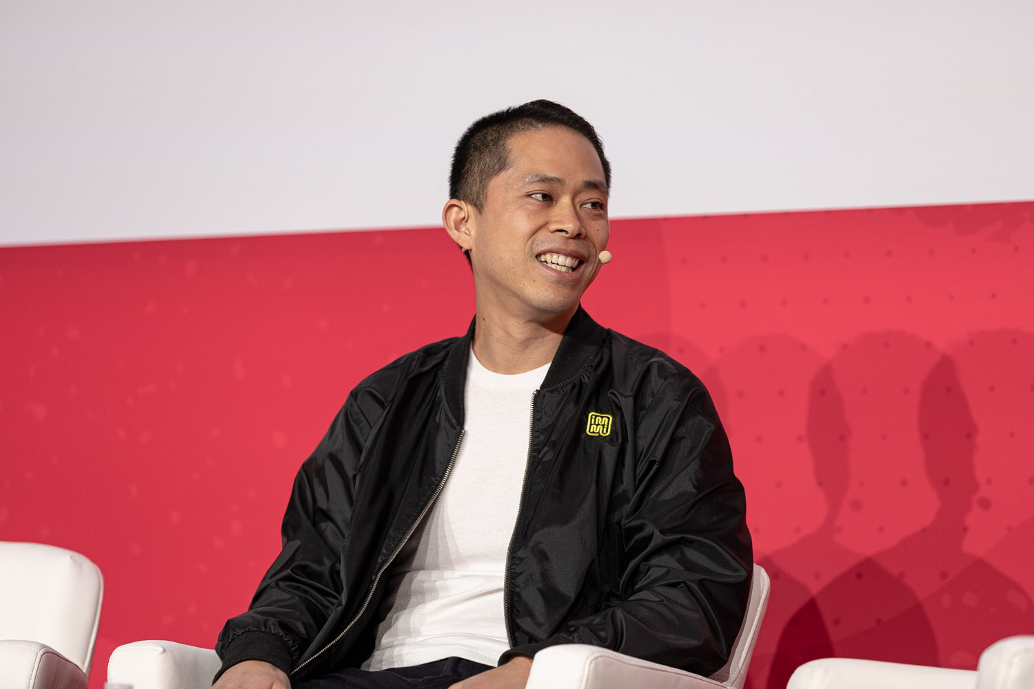 A smiling man in a black jacket seated on stage.