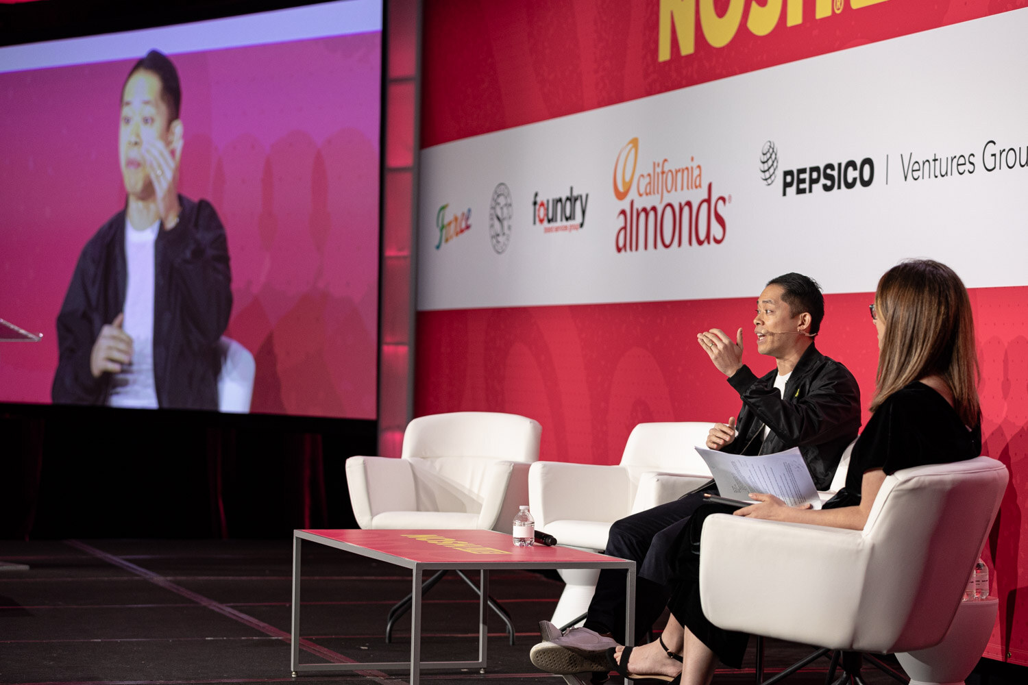 Two people seated on stage with a man shown on a large screen behind them.