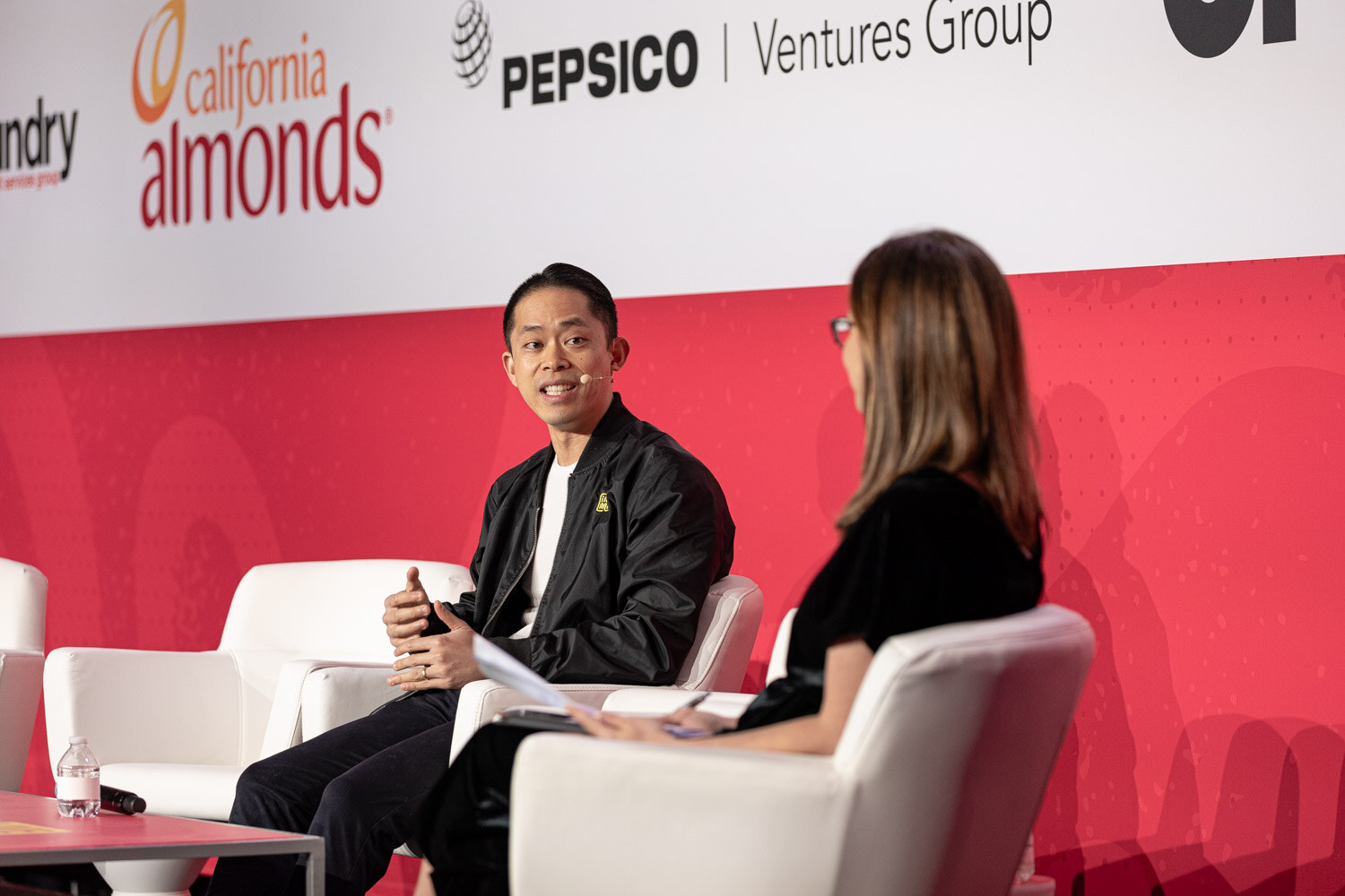 A woman and a man seated in white chairs in front of California Almonds and Pepsico Ventures Group signs.