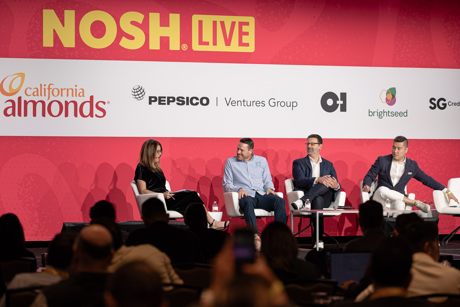 Four people seated on stage with the Nosh Live backdrop and a speaker on the screen.