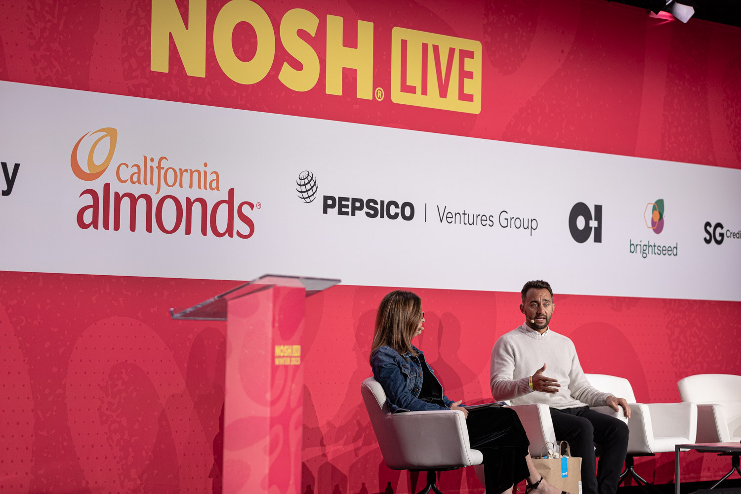 A woman and a man seated on stage next to a podium in front of the Nosh Live backdrop.