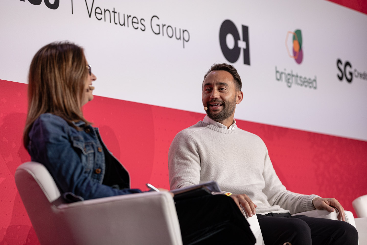 A man in a white sweater talking with a woman in a denim jacket in white chairs.
