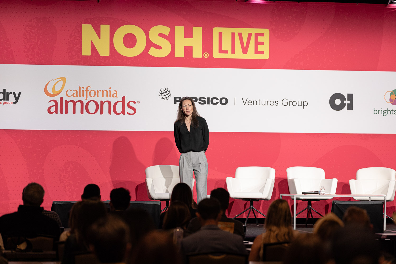 A woman in black standing alone on a stage in front of the NOSH LIVE backdrop.