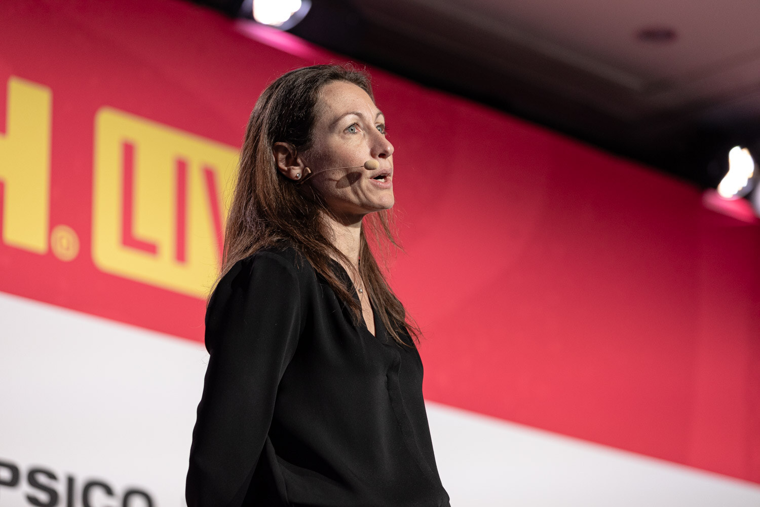 A woman with long hair and a headset microphone speaking on stage.