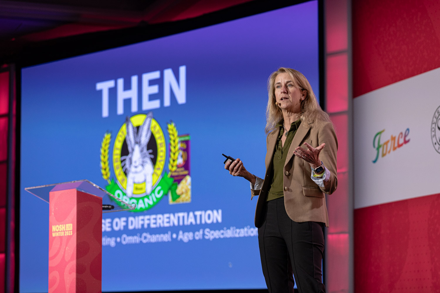 A woman in a tan blazer standing on stage next to a blue slide reading Then.