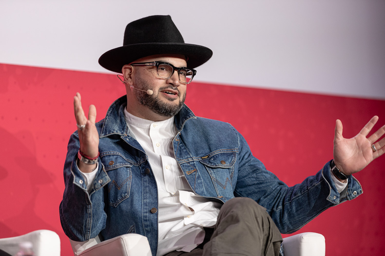 A man in a black hat and denim jacket gesturing while seated on stage.