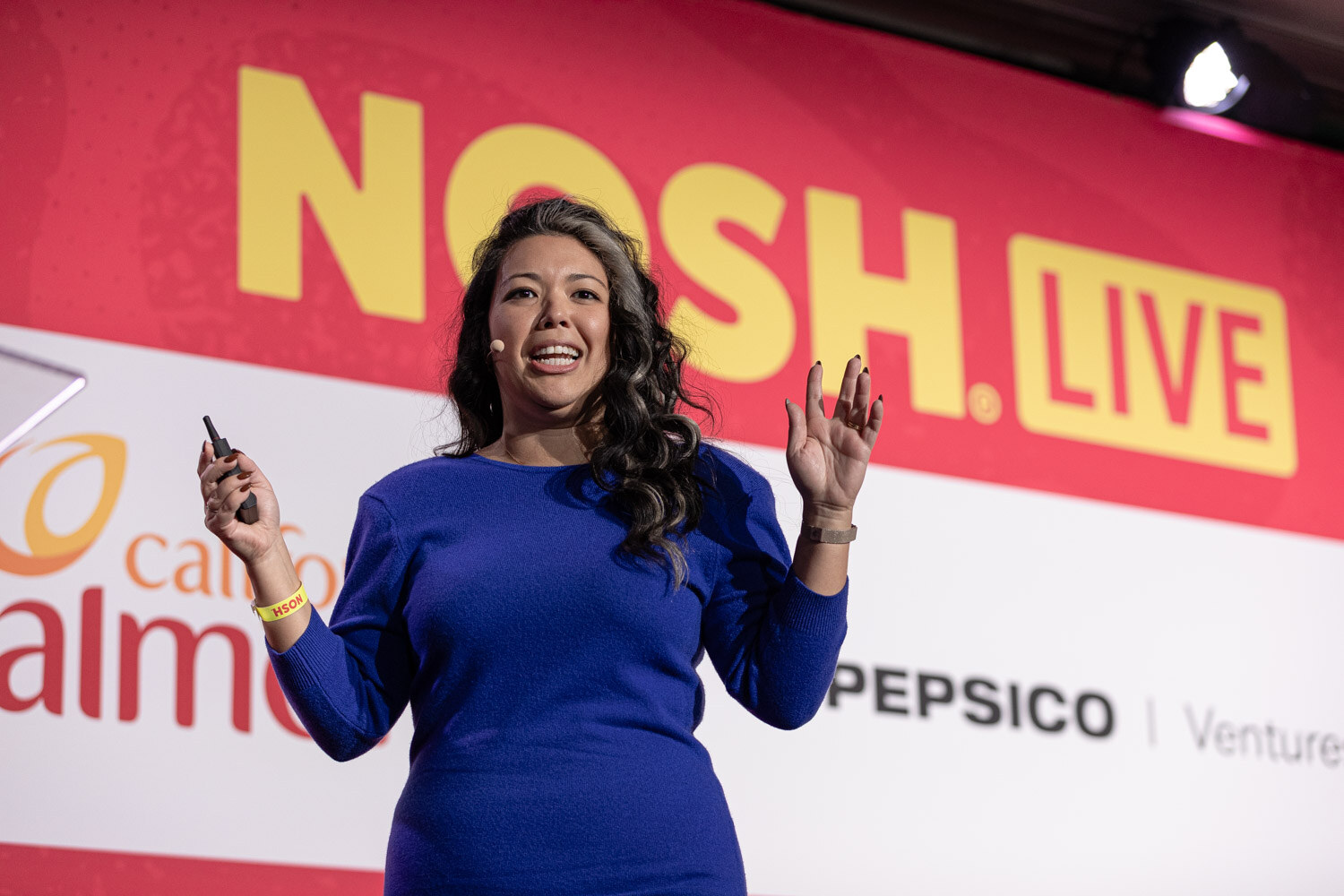 A woman in a blue dress holding a clicker and gesturing in front of the Nosh Live backdrop.