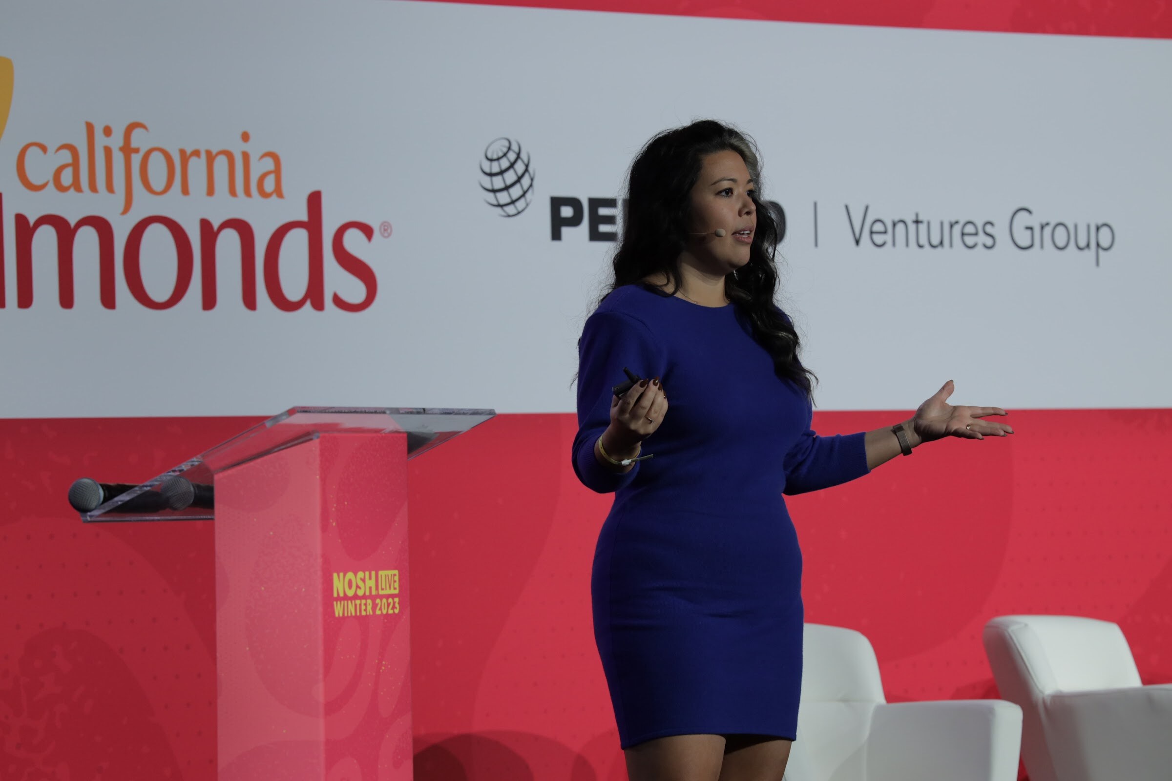 A woman in a blue dress speaking on a stage in front of a California Almonds banner.