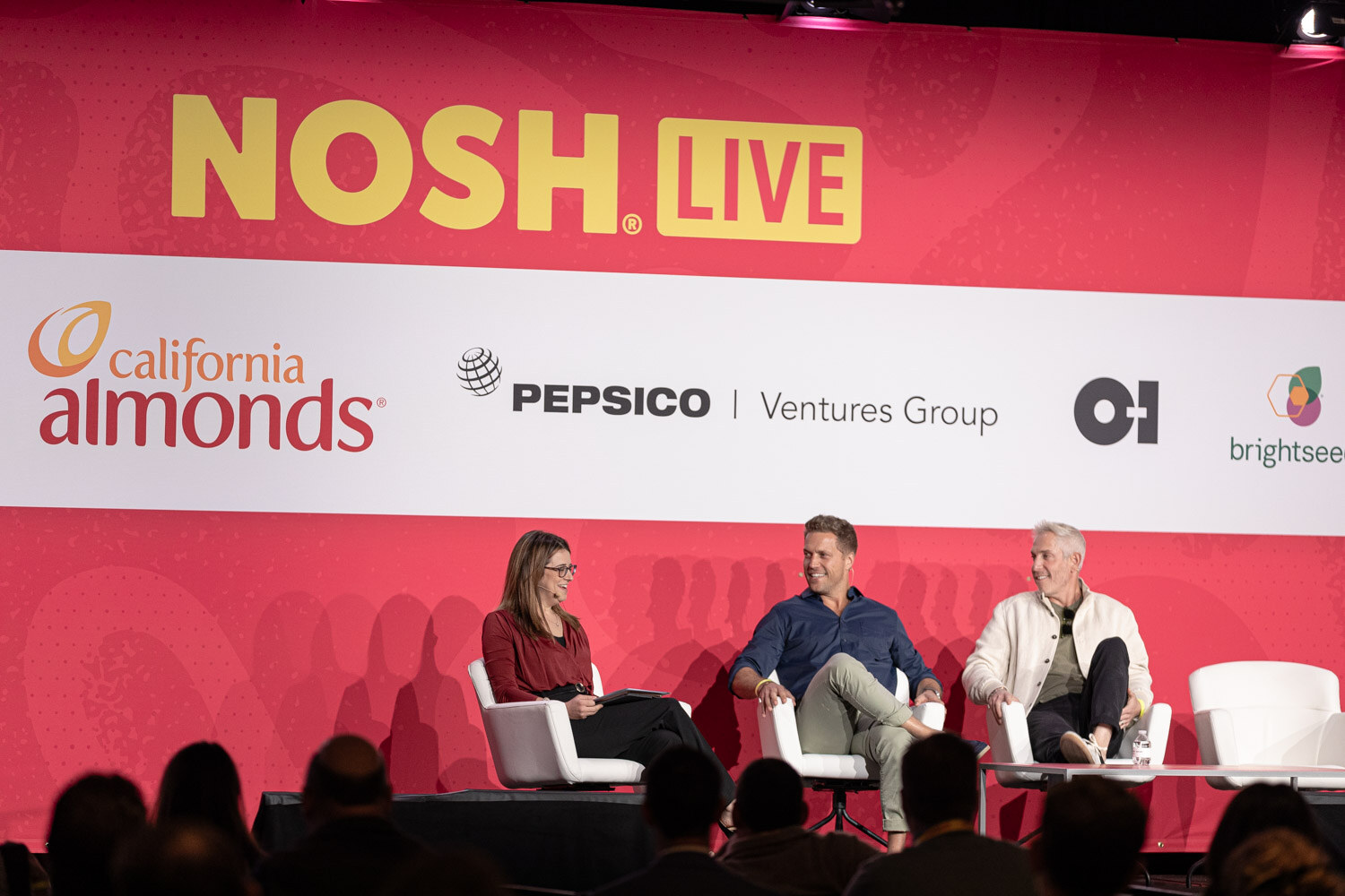 Three people seated on stage in front of the Nosh Live backdrop.