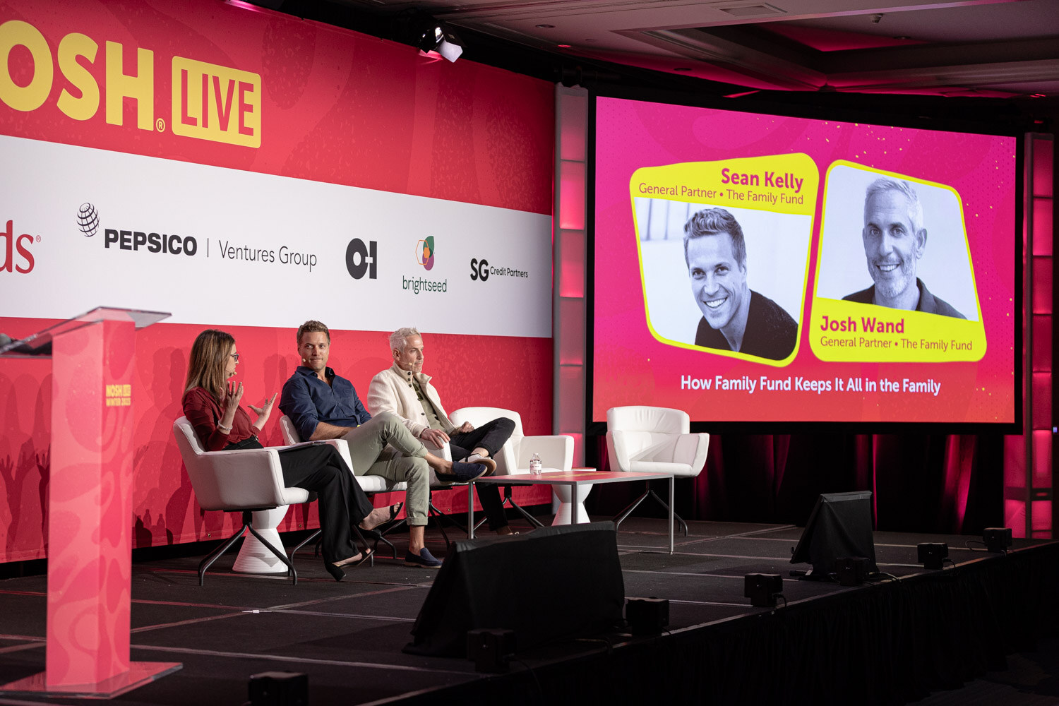 A stage with three people seated and a screen showing two speakers and the session title.