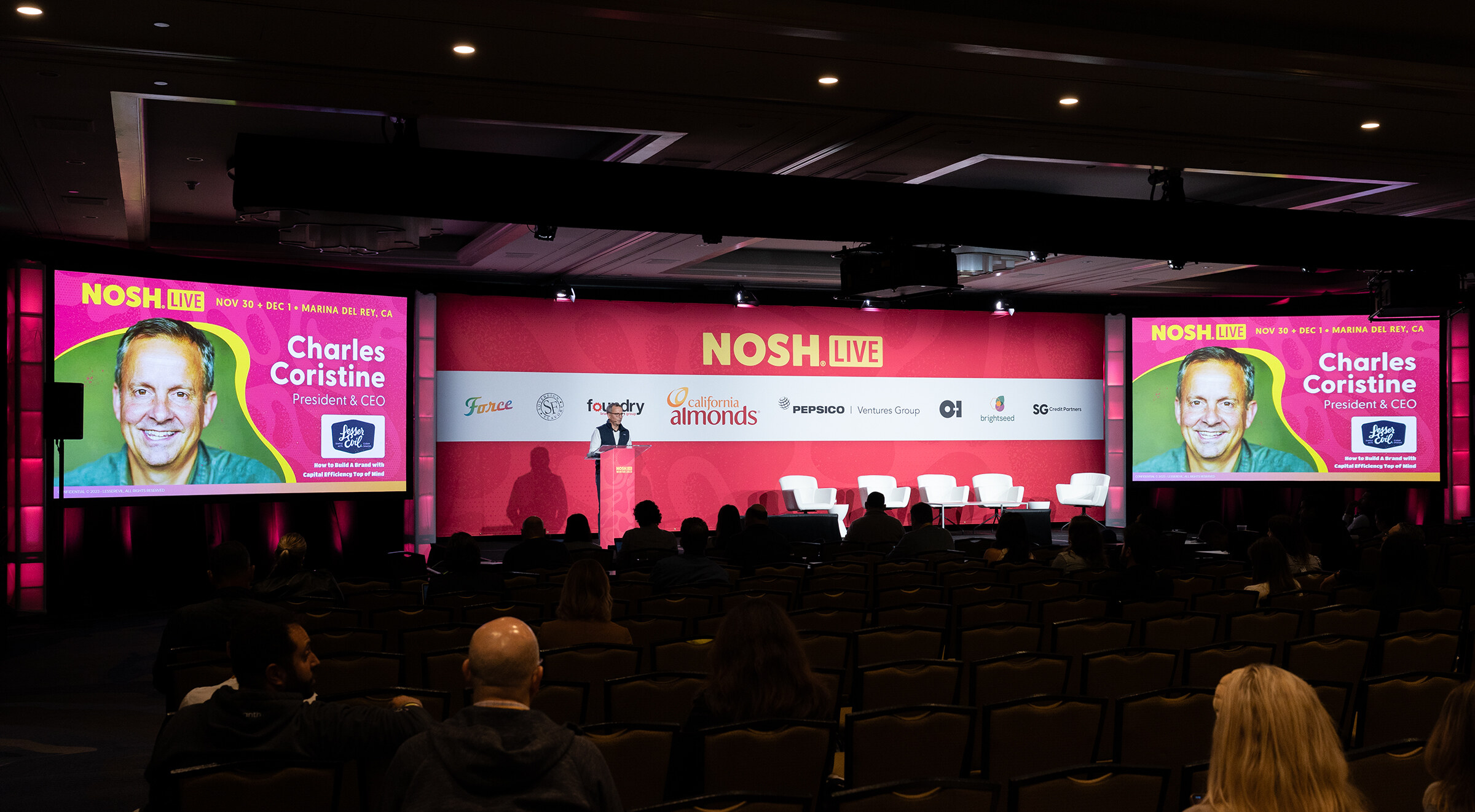 A stage with a man at a podium and large screens showing a speaker's name and photo.