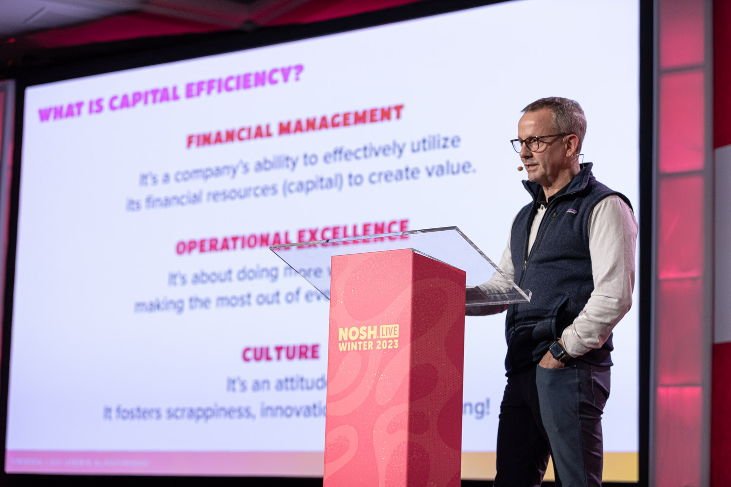 A man in a navy vest standing at a podium next to a slide titled What Is Capital Efficiency.