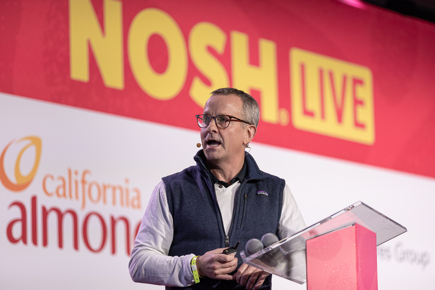 A man with glasses in a navy vest at a podium in front of the Nosh Live backdrop.