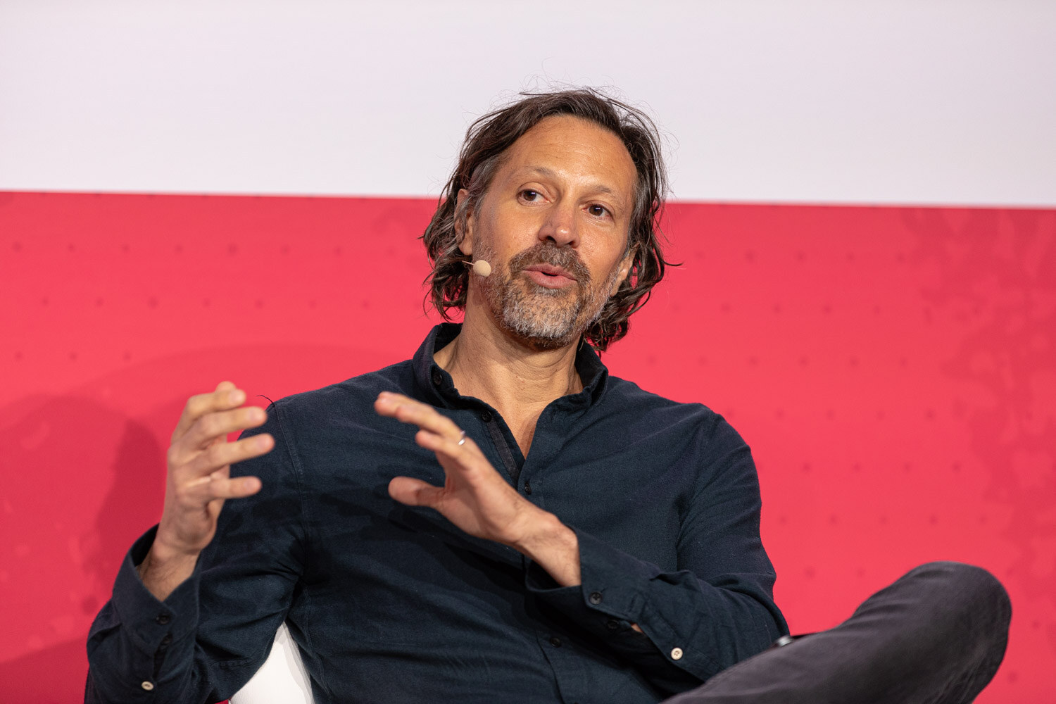 A man with wavy hair in a dark shirt gesturing while seated on stage.