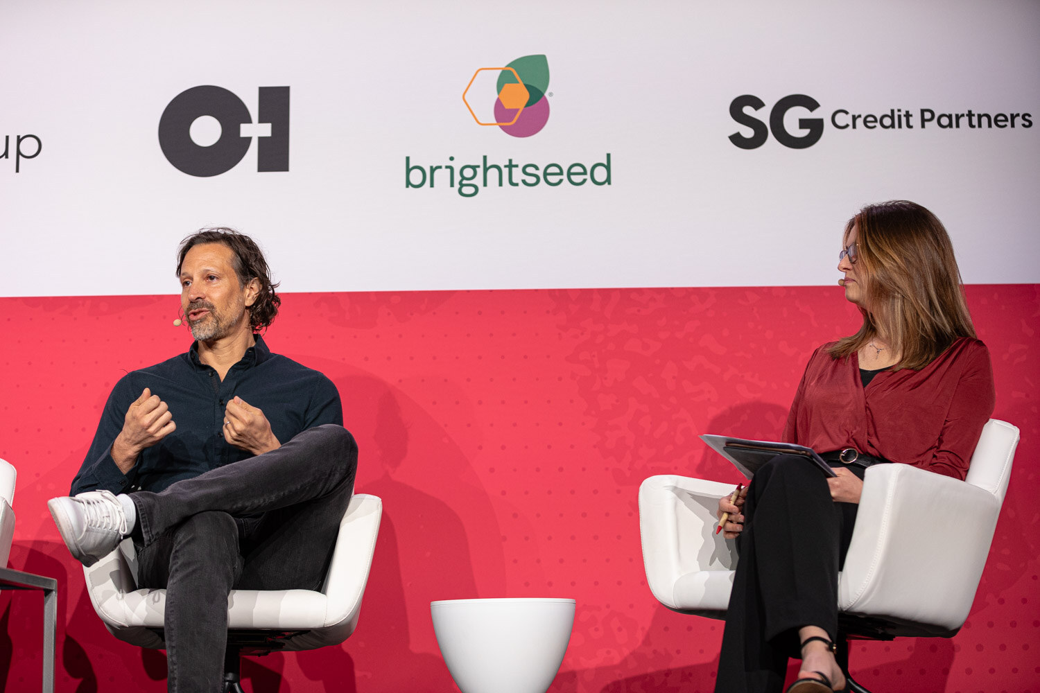 A woman in a red top and a man in a dark shirt seated in white chairs in front of brightseed and SG Credit Partners signs.
