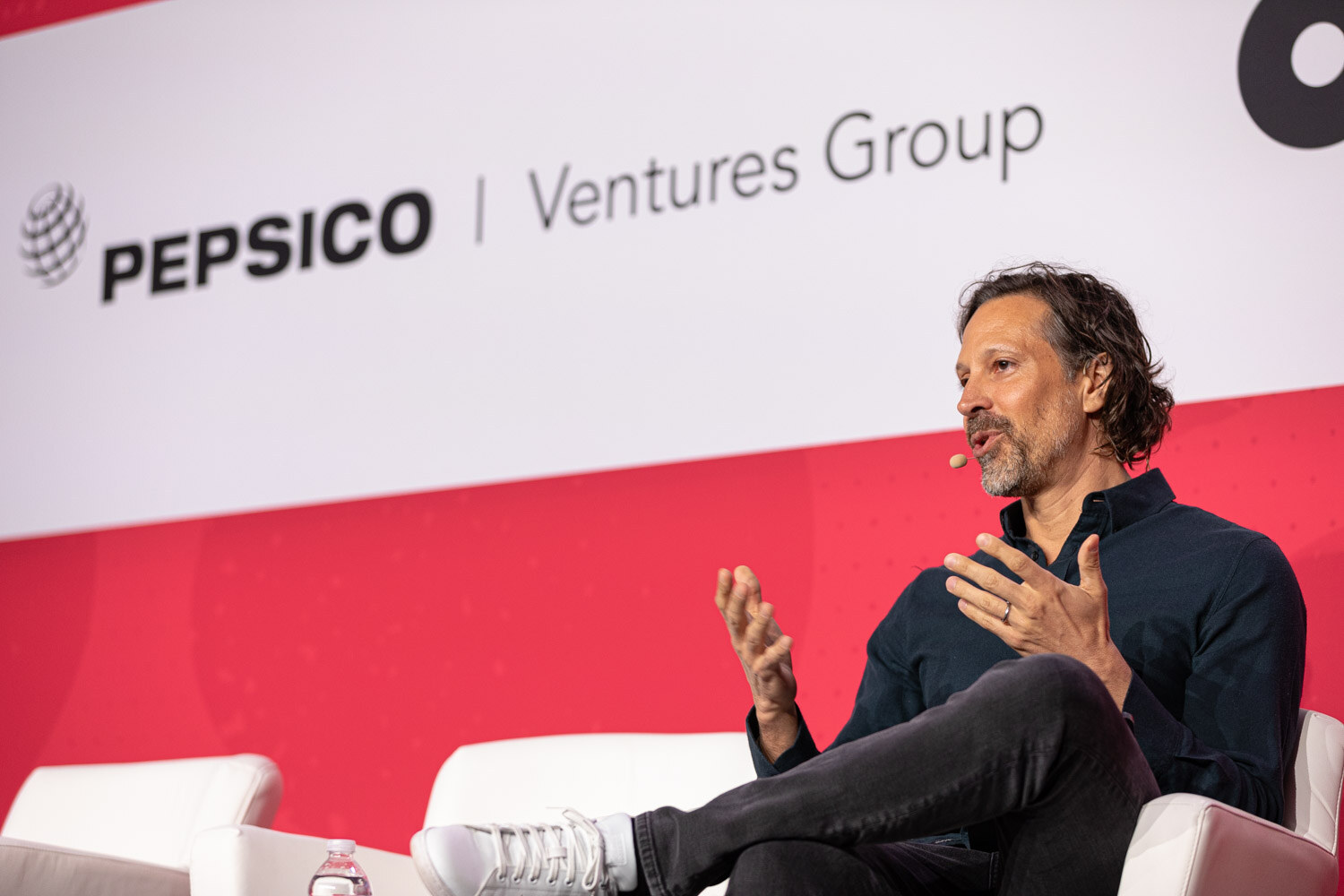 A man with wavy hair gesturing while seated in front of a Pepsico Ventures Group sign.