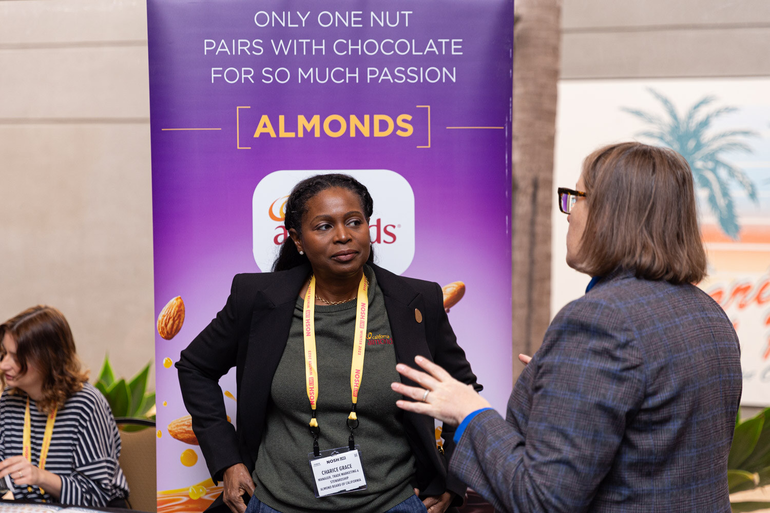 A woman in a blazer speaks with another attendee in front of a purple Almonds banner.