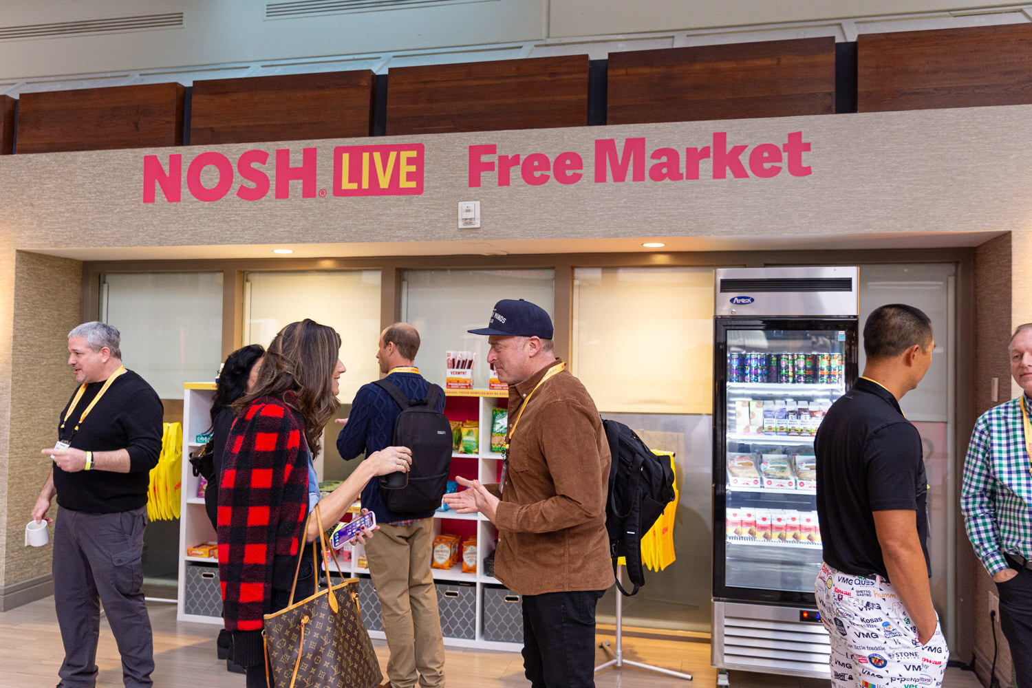 People standing in front of shelves and a refrigerator under a sign reading NOSH LIVE Free Market.