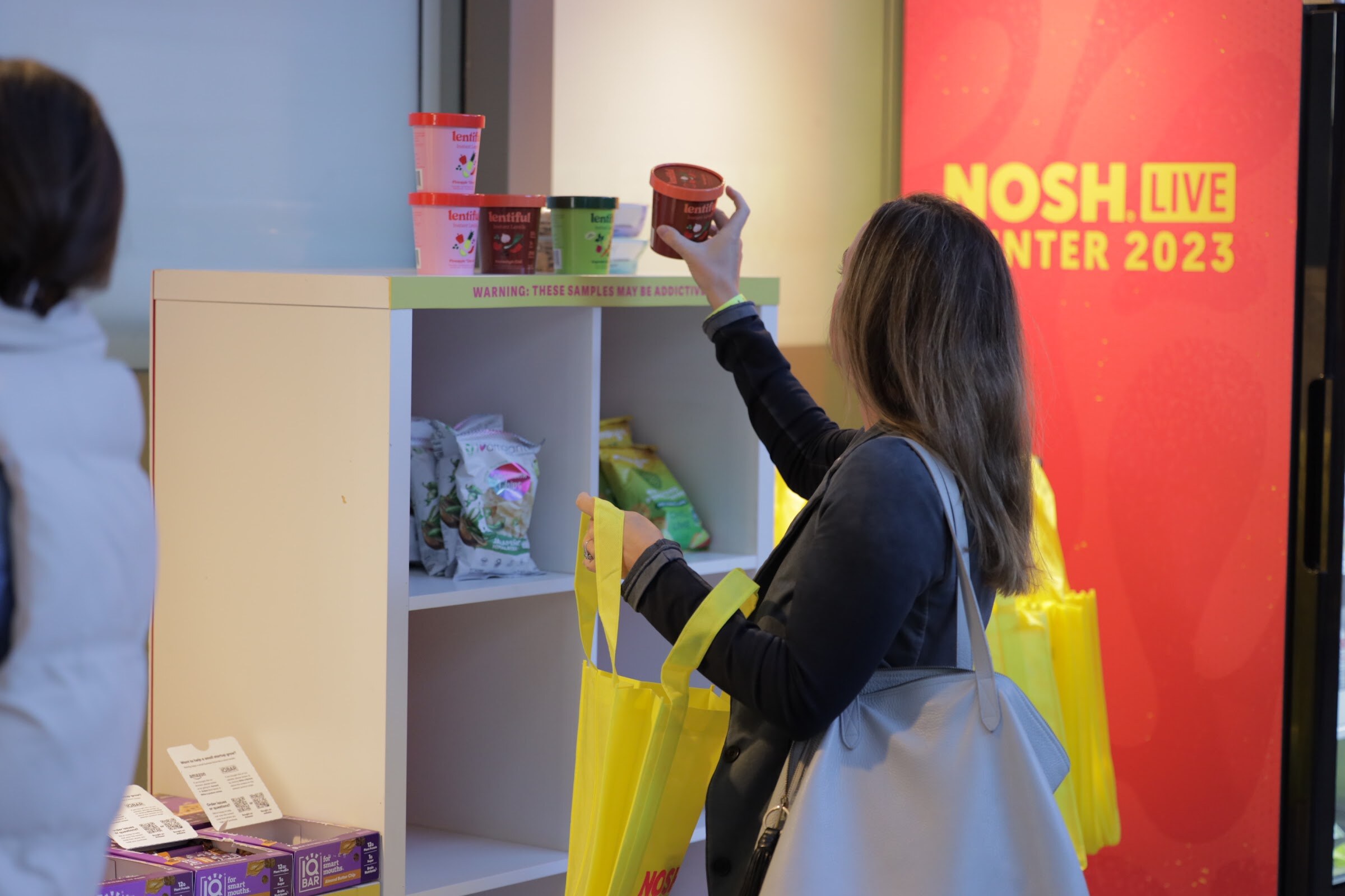 An attendee with a tote bag reaching toward shelves of snack bags beside a NOSH LIVE Winter 2023 panel.