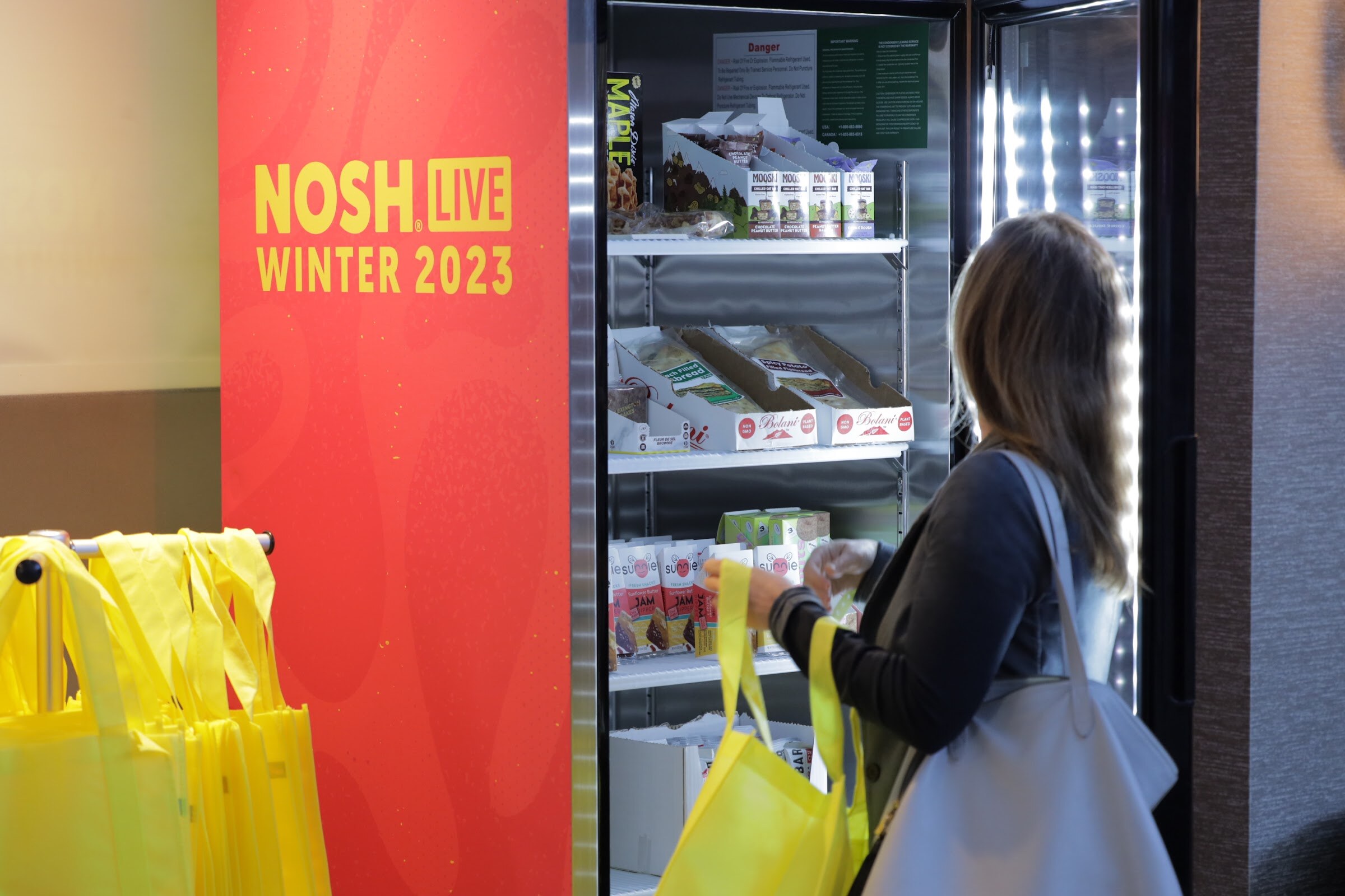 A woman with a tote bag looks into a lit refrigerator stocked with packaged food samples.