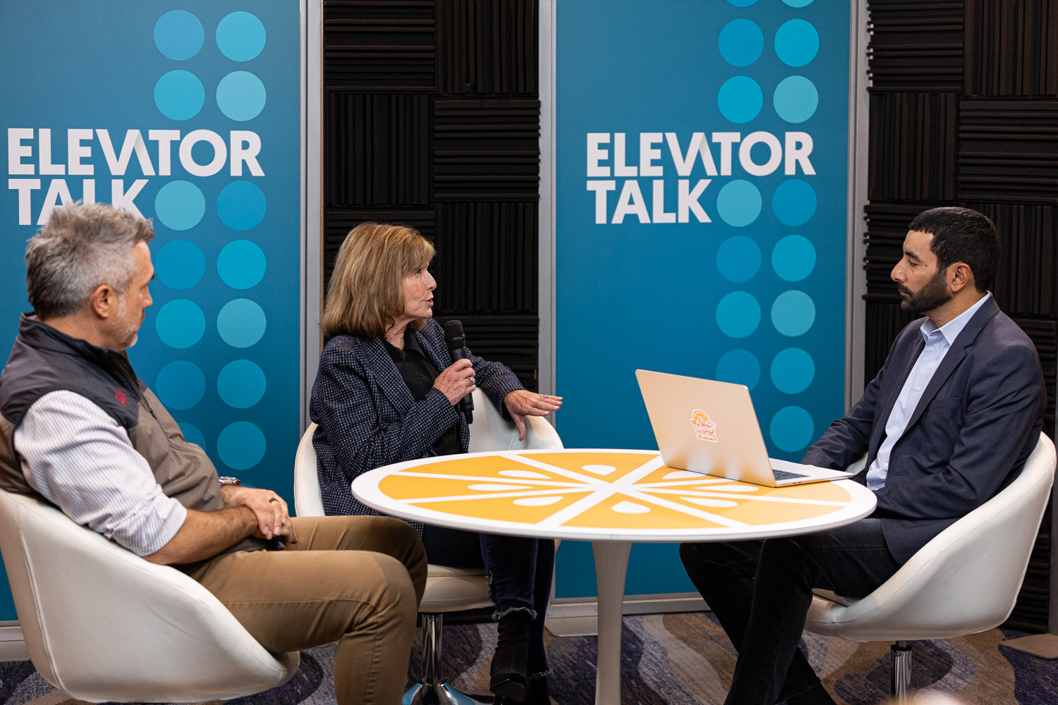 A host and two guests seated around a round table in front of Elevator Talk banners.