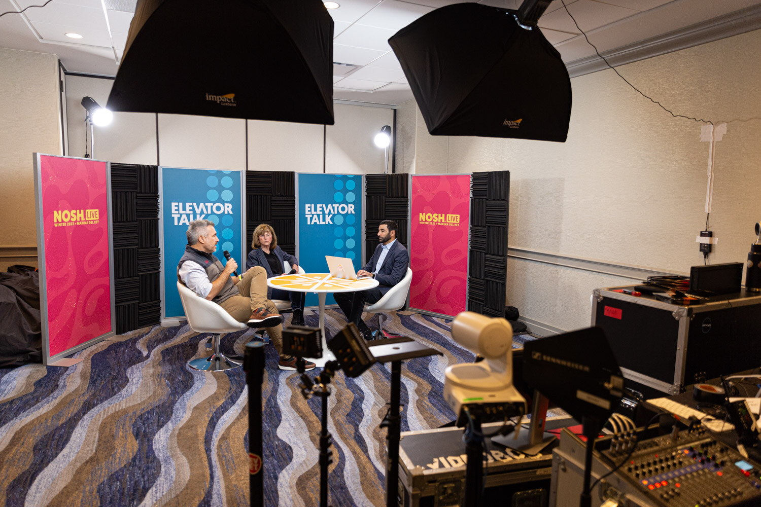 A wide view of a small studio with lights, cameras and an Elevator Talk backdrop.