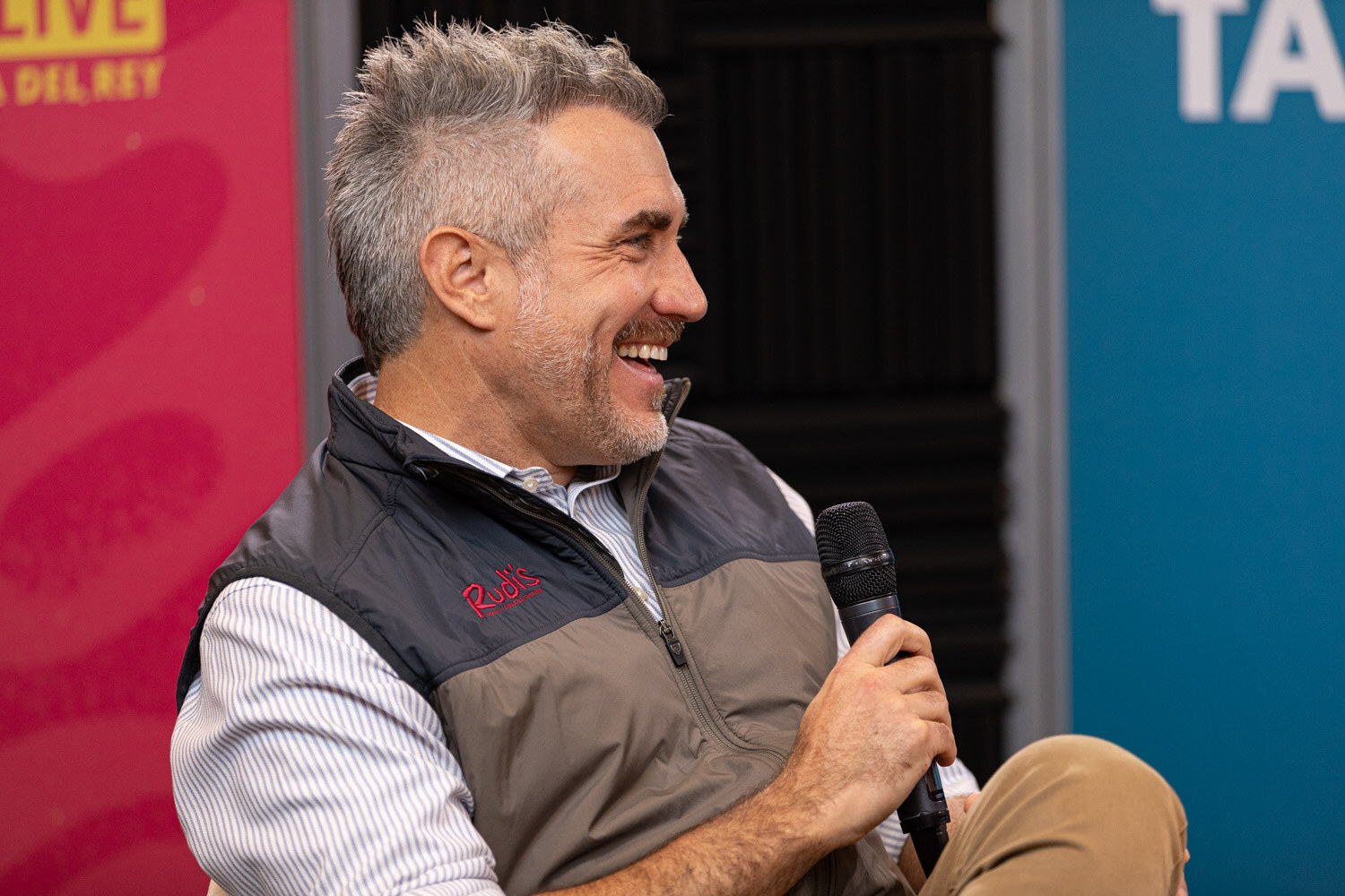 A smiling man in a vest holding a microphone in front of Elevator Talk and Nosh Live banners.