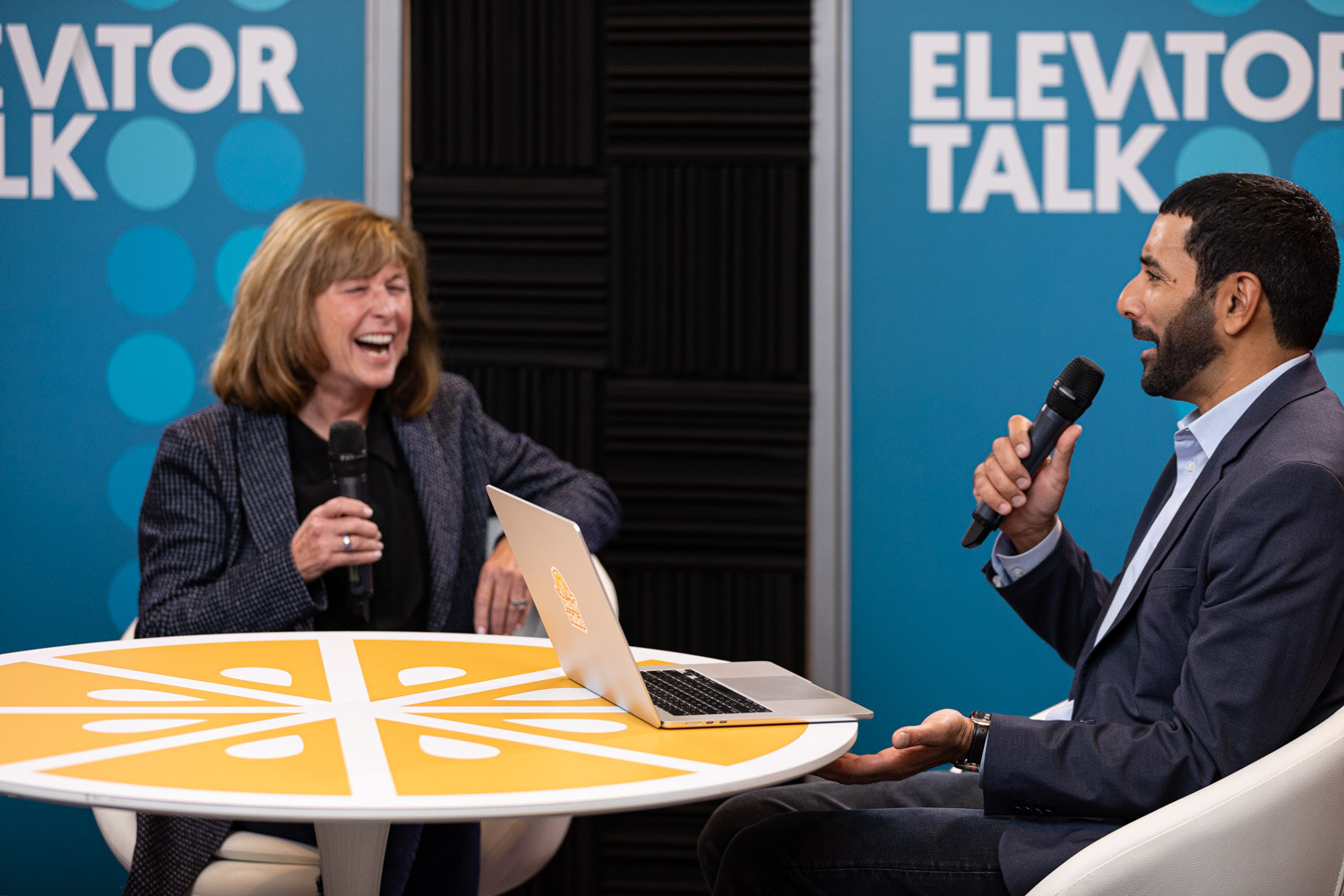 A woman laughing with a microphone seated across from a man in a suit holding a microphone.
