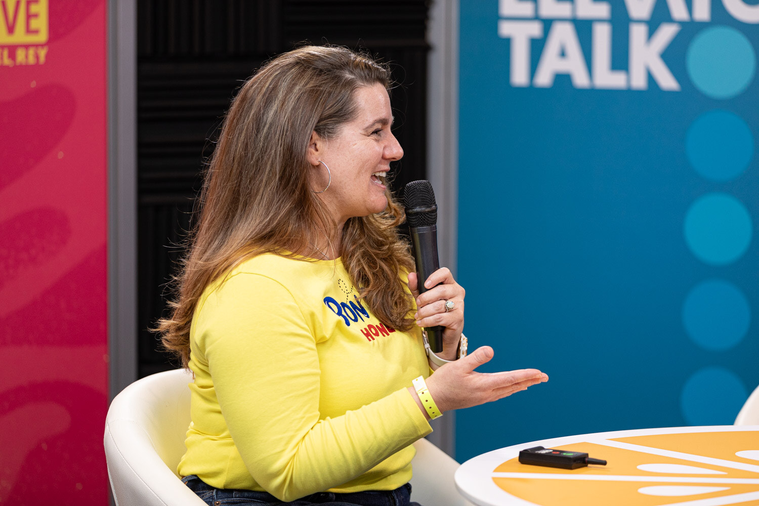 A woman in a yellow sweater speaking into a microphone in front of an Elevator Talk banner.
