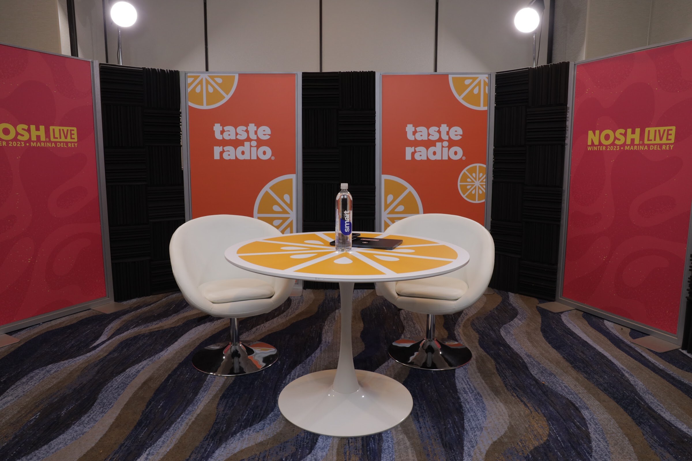 An empty studio with two white chairs and an orange table between Taste Radio banners.