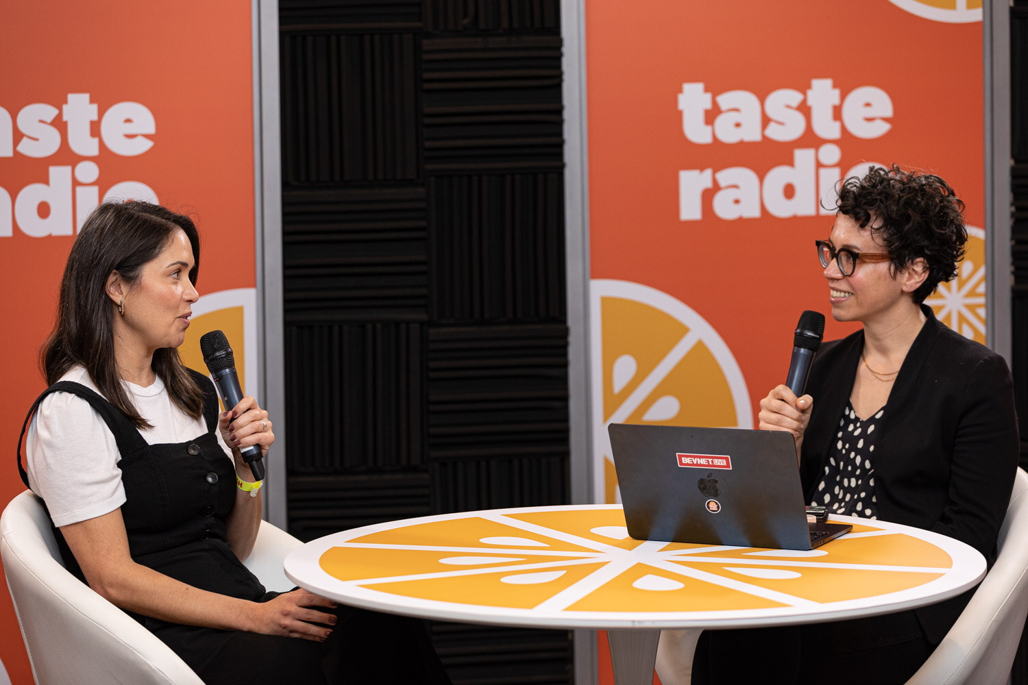 Two women at a laptop seated at a round table in front of a Taste Radio banner.