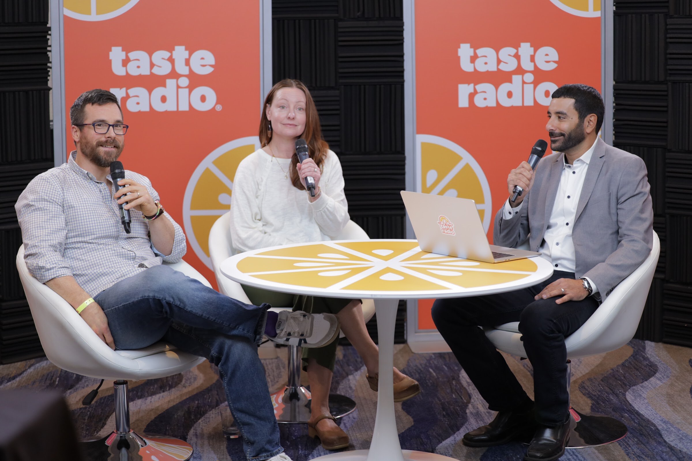 Three people with microphones seated around a table in front of orange Taste Radio banners.