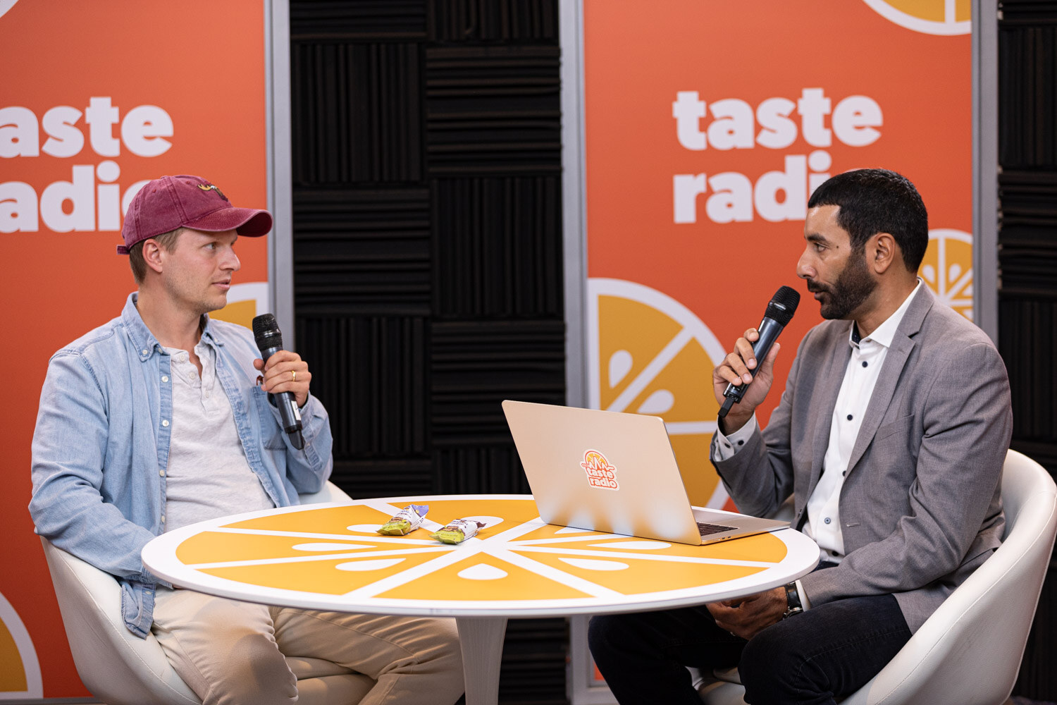A man in a maroon cap talking with a host holding a microphone at the Taste Radio table.