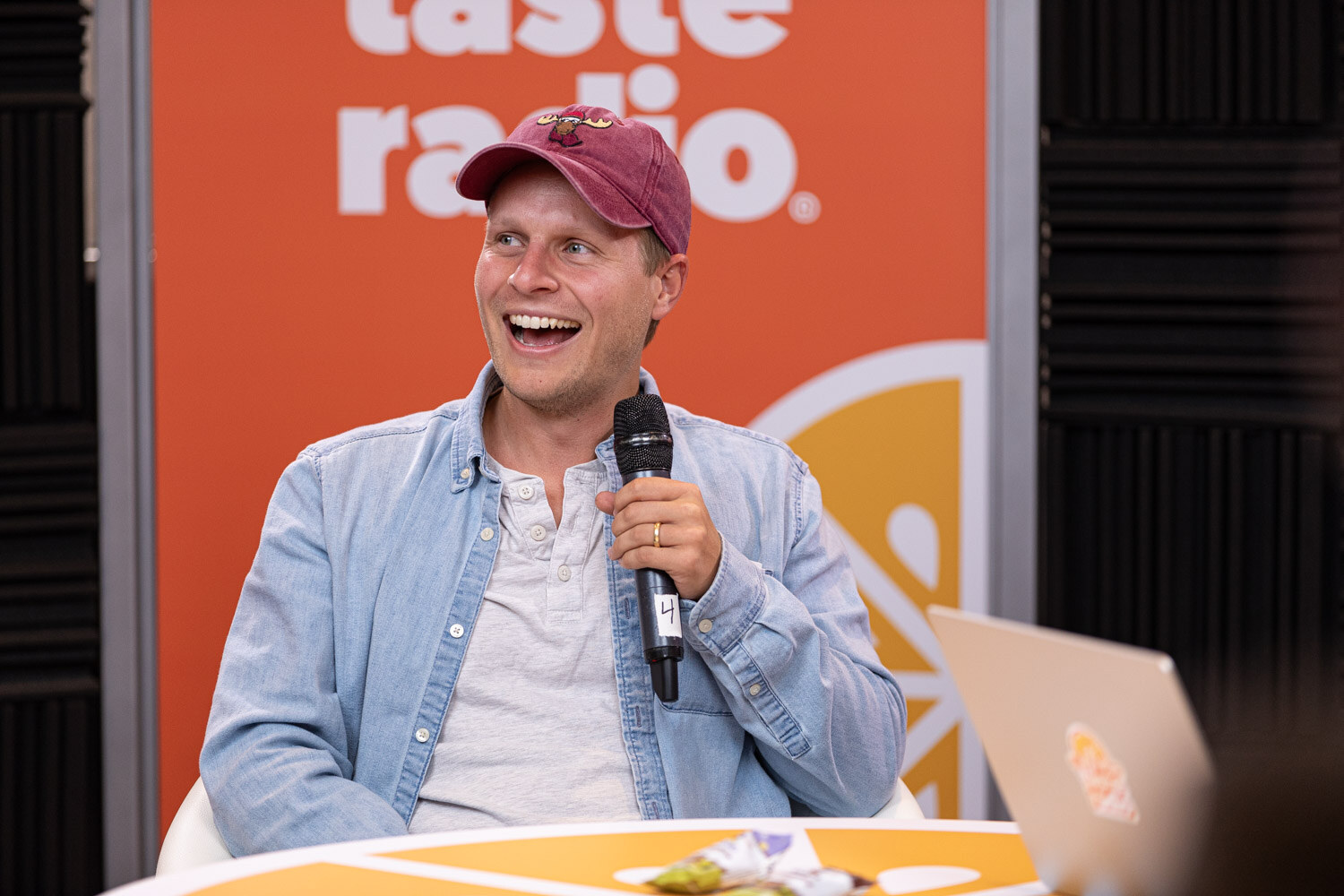 A smiling man in a denim shirt and maroon cap holding a microphone.