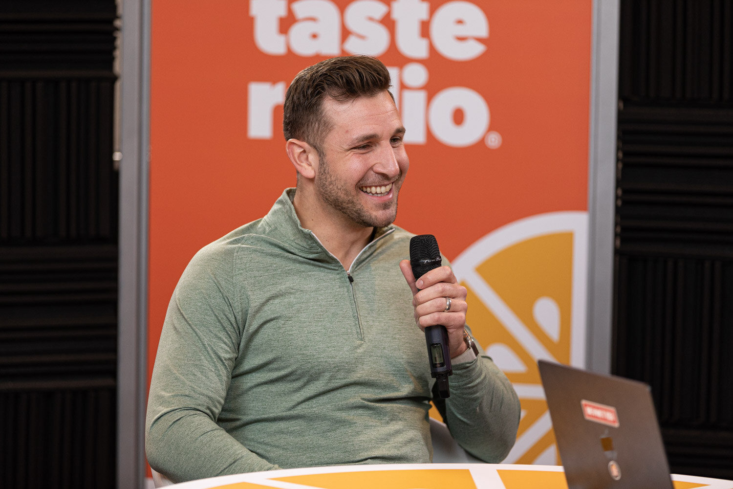 A smiling man in a green quarter-zip pullover holding a microphone in front of a Taste Radio banner.