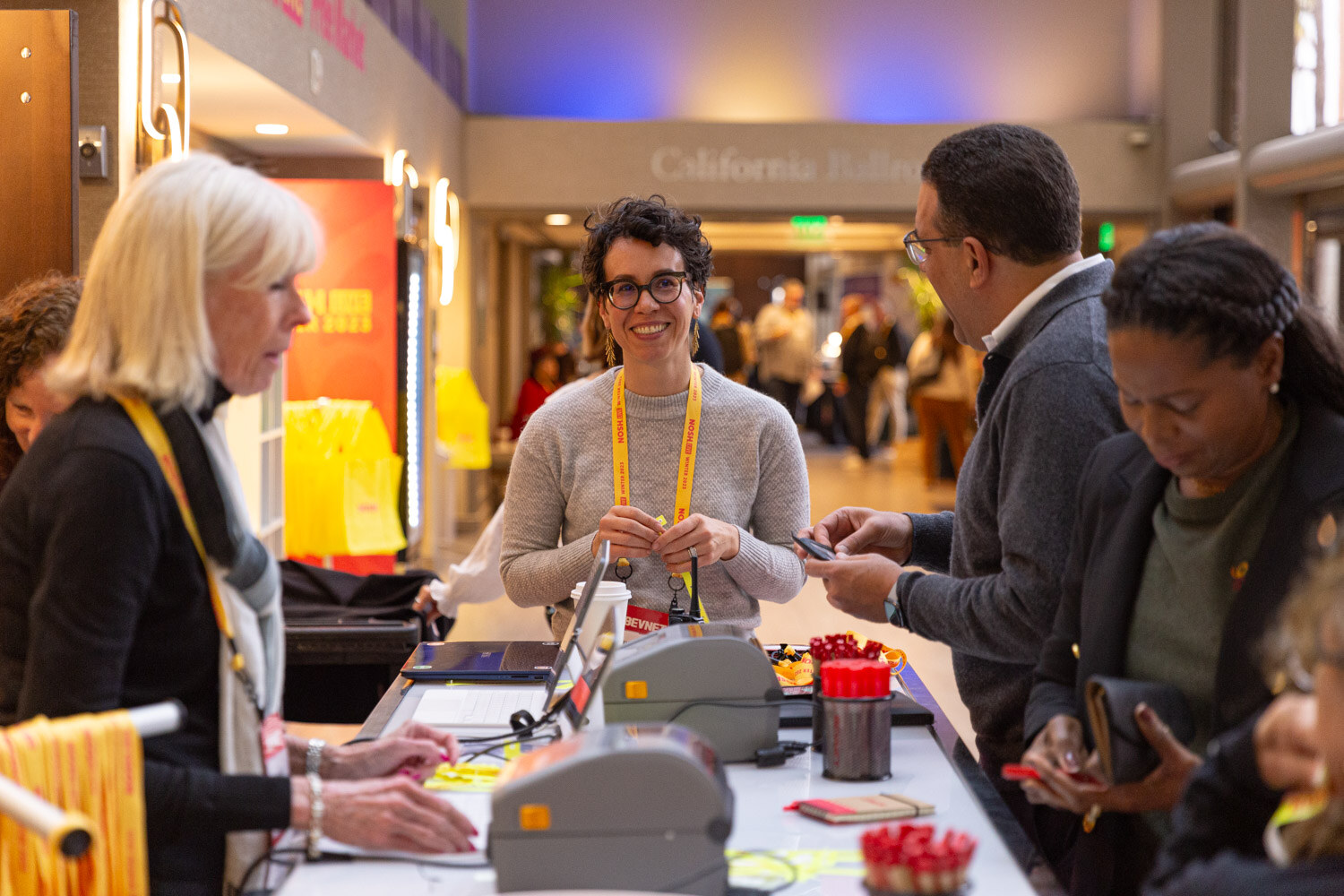 Staff at a registration counter with label printers hand badges to arriving attendees.