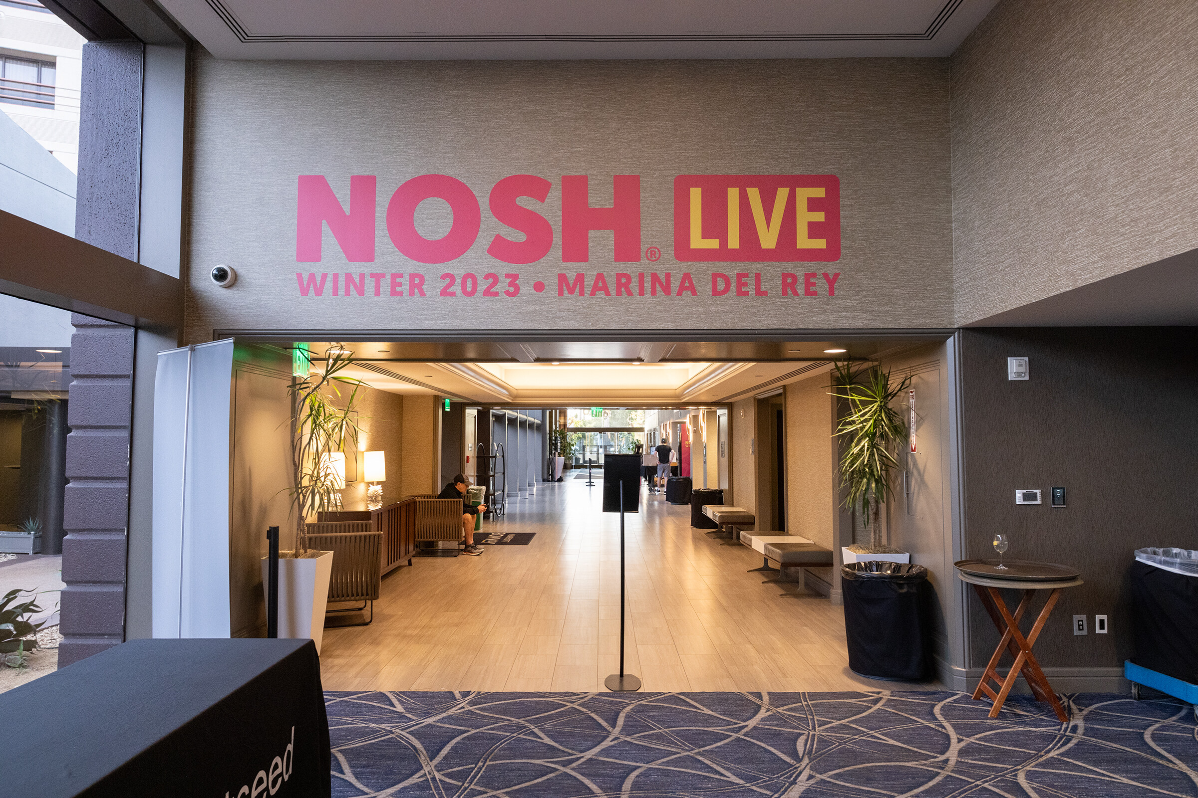 A pink NOSH LIVE Winter 2023 Marina Del Rey sign on a wall above a hotel hallway.