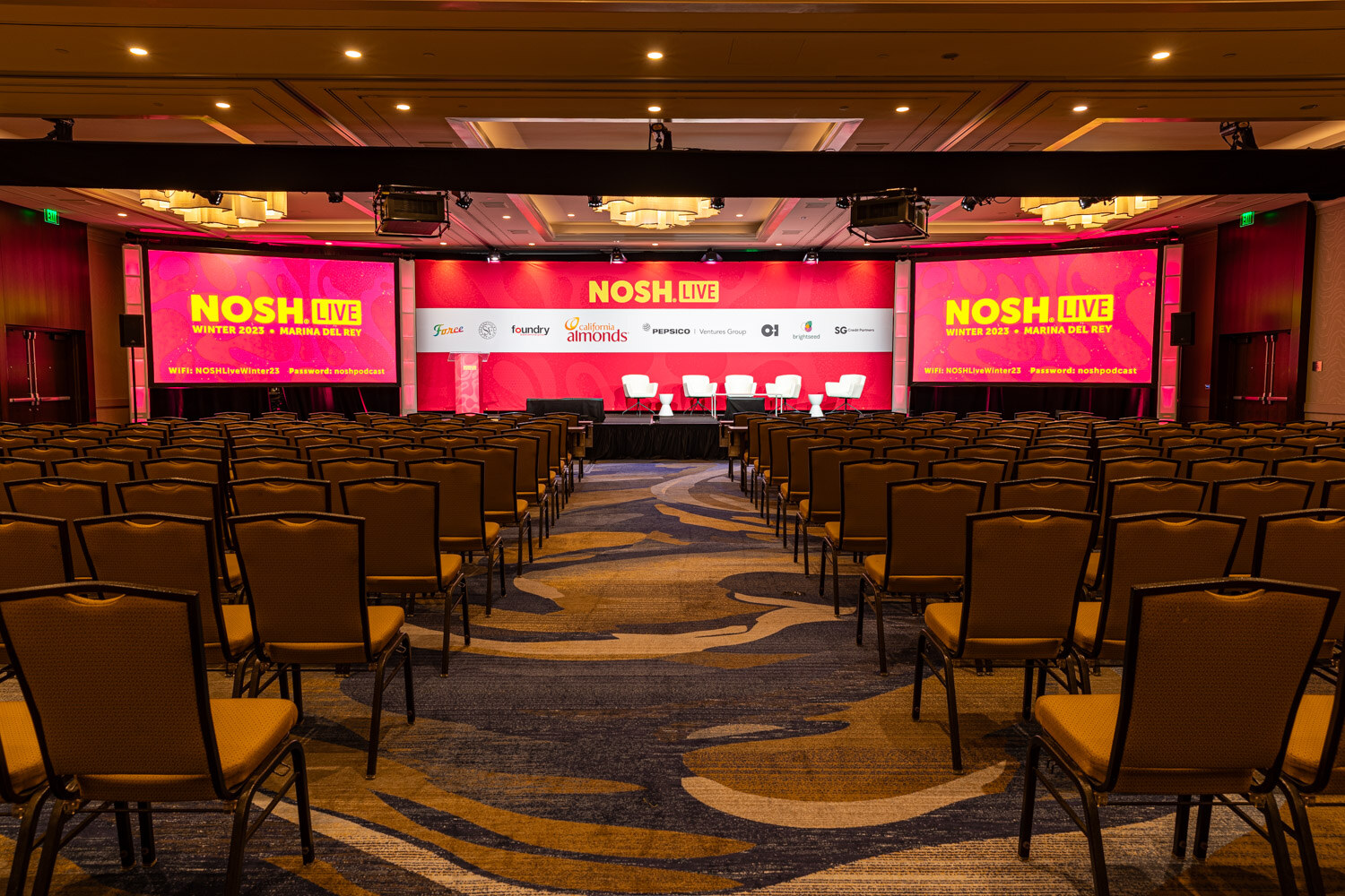 An empty ballroom with rows of chairs facing a pink stage backdrop and screens reading NOSH LIVE.