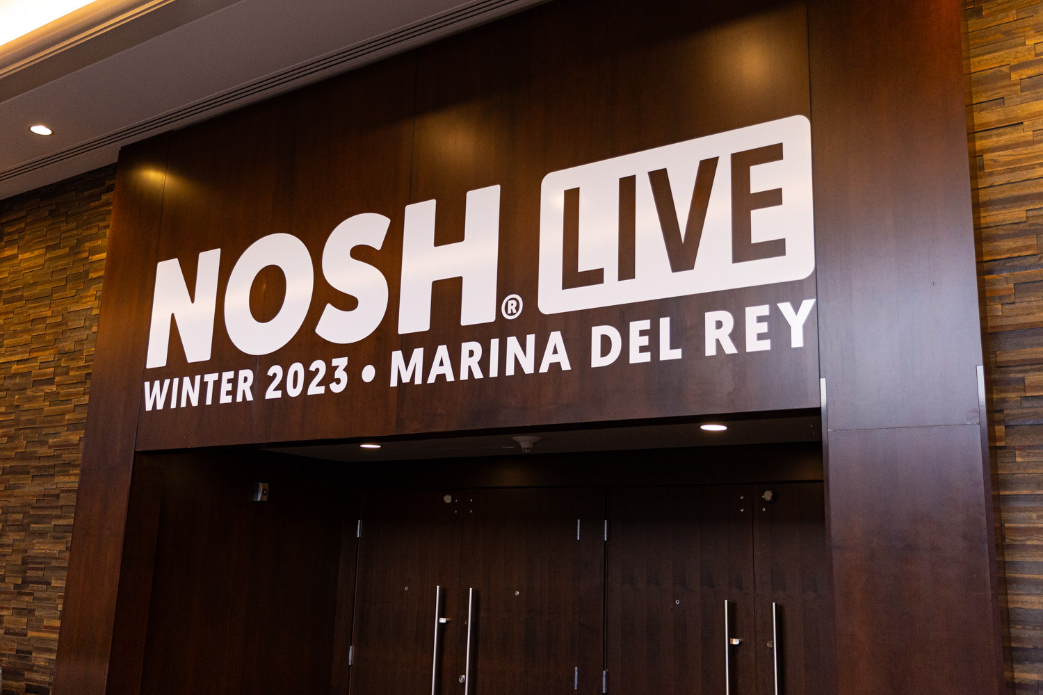 White NOSH LIVE Winter 2023 Marina Del Rey lettering on a dark wood wall above double doors.