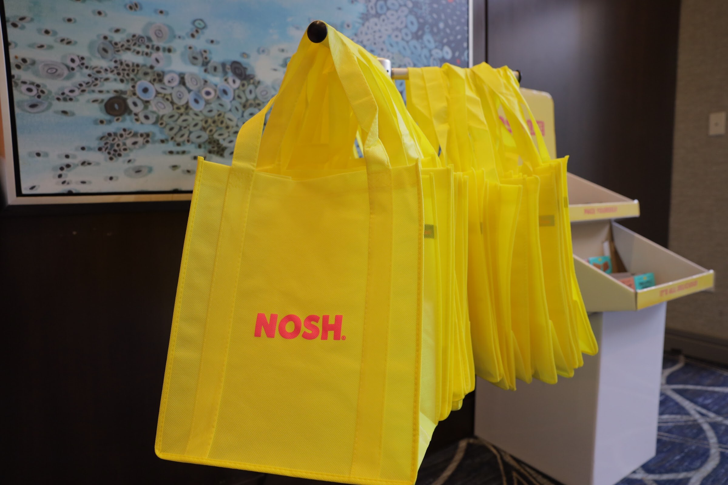 Yellow tote bags printed with NOSH hanging next to cardboard display shelves.