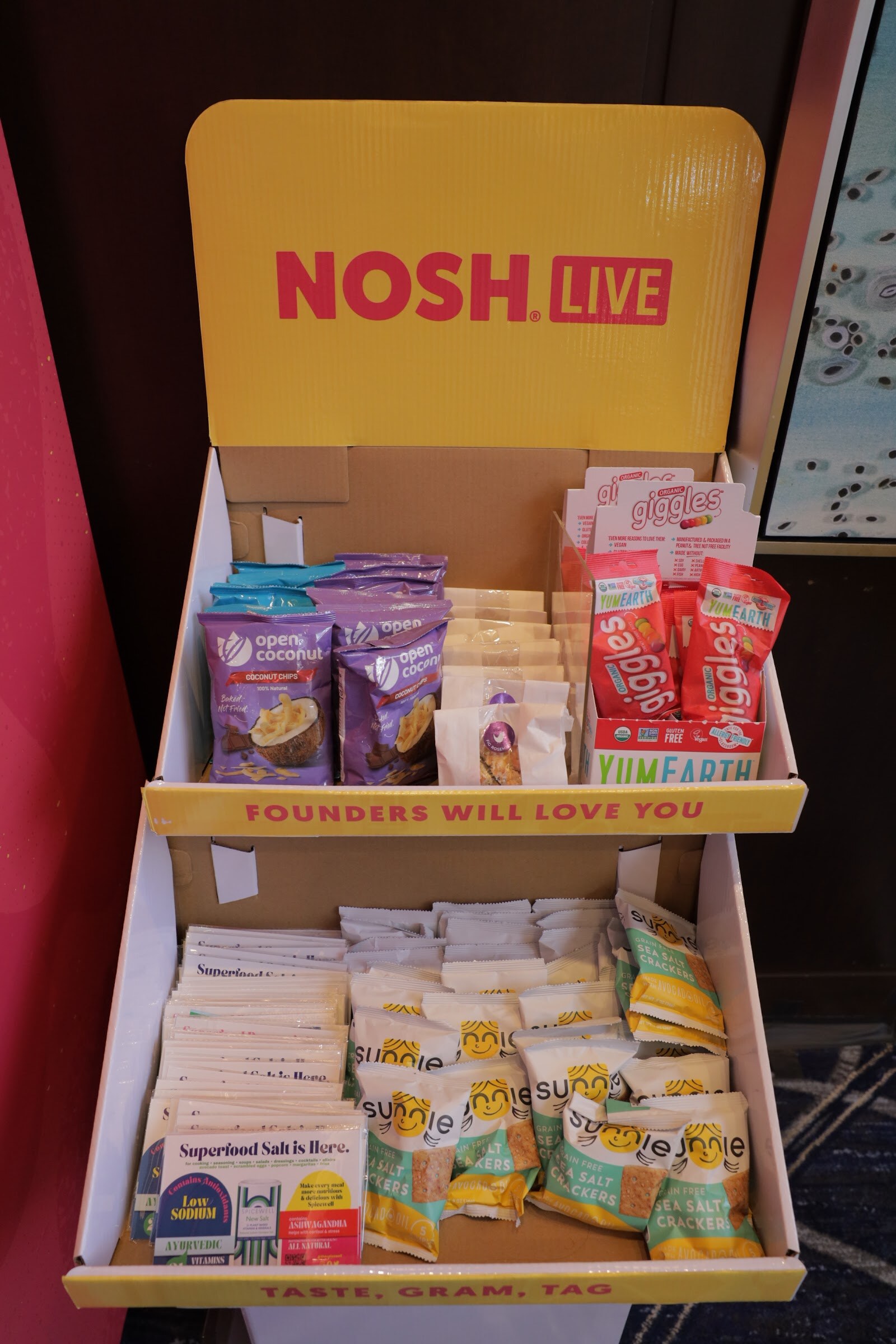 A yellow NOSH LIVE cardboard display stocked with packaged snack samples.