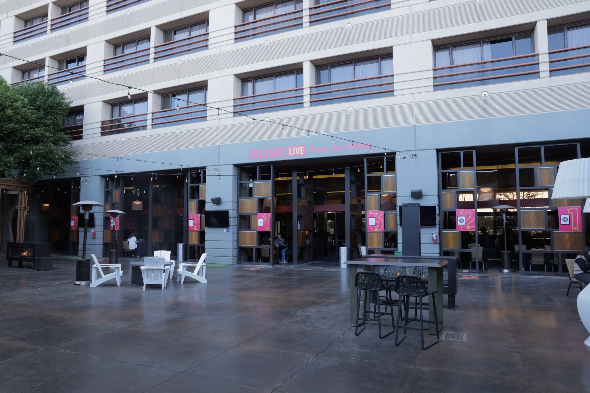 A hotel courtyard with white chairs and high tables in front of a glass-fronted building.