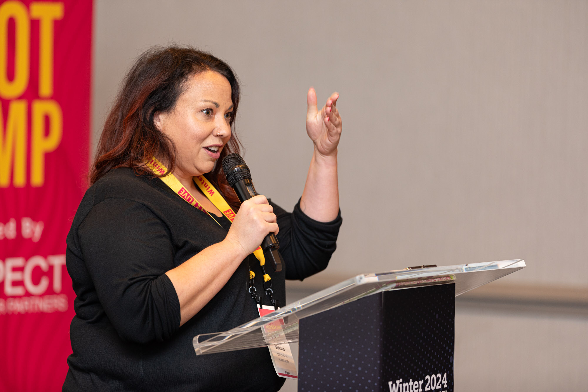 A woman with auburn hair and a lanyard speaking at a clear podium beside a red Nosh Live Boot Camp banner.